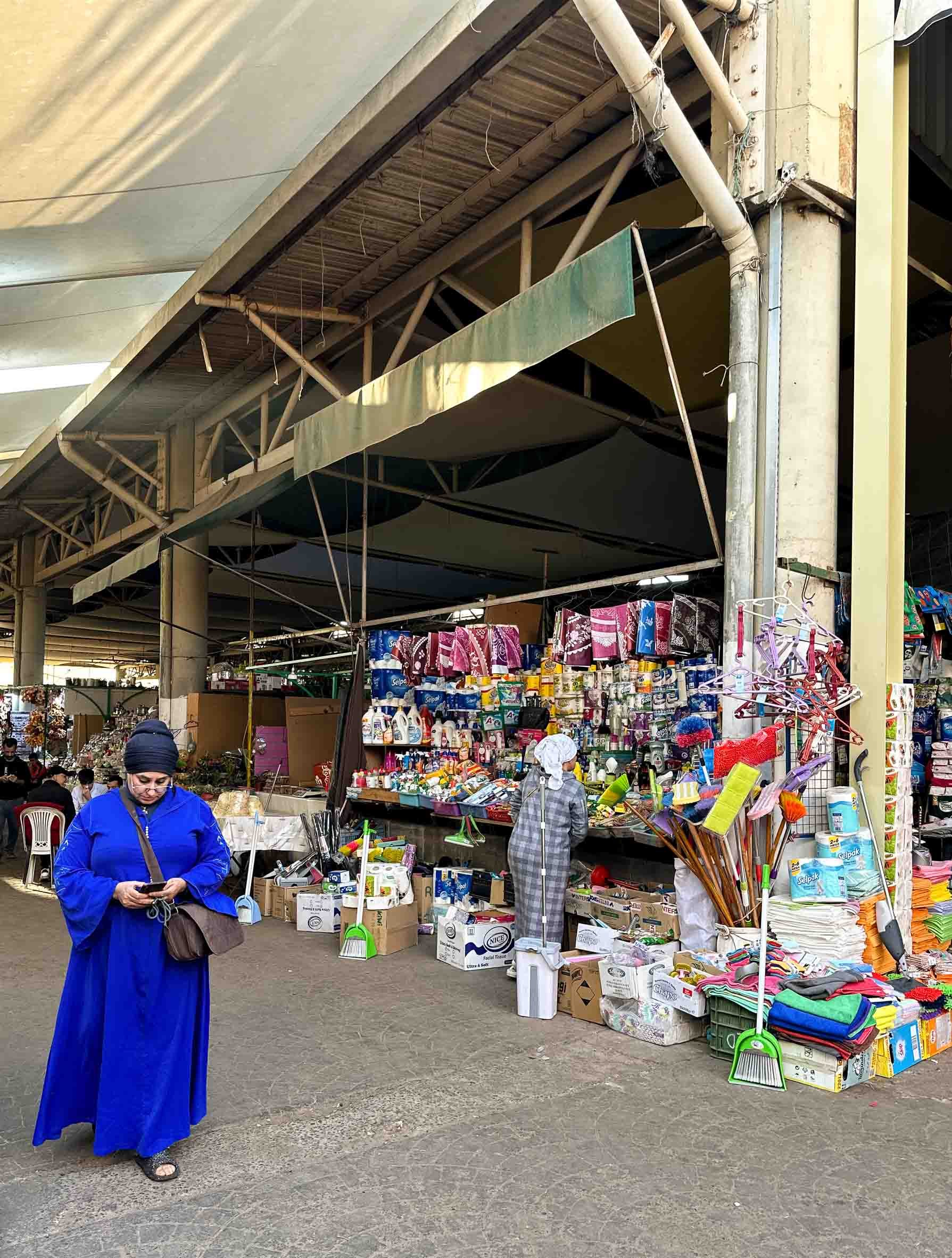 Street market stall with household items, cleaning supplies, and textiles, and a woman in a blue dress looking at her phone.