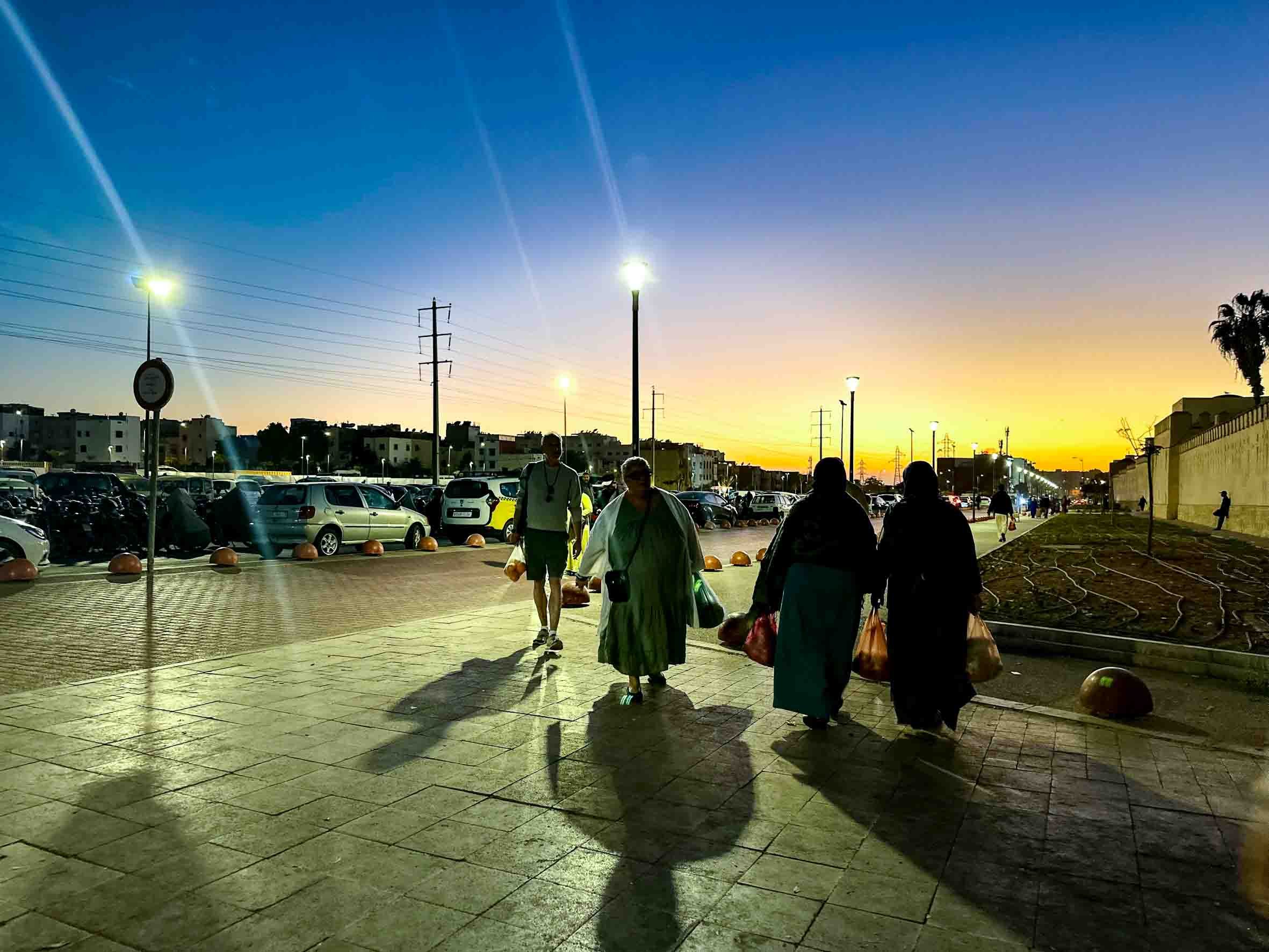 People walking on a sidewalk at sunset, carrying shopping bags, with a parking lot and cars on the left and a building and palm trees on the right.