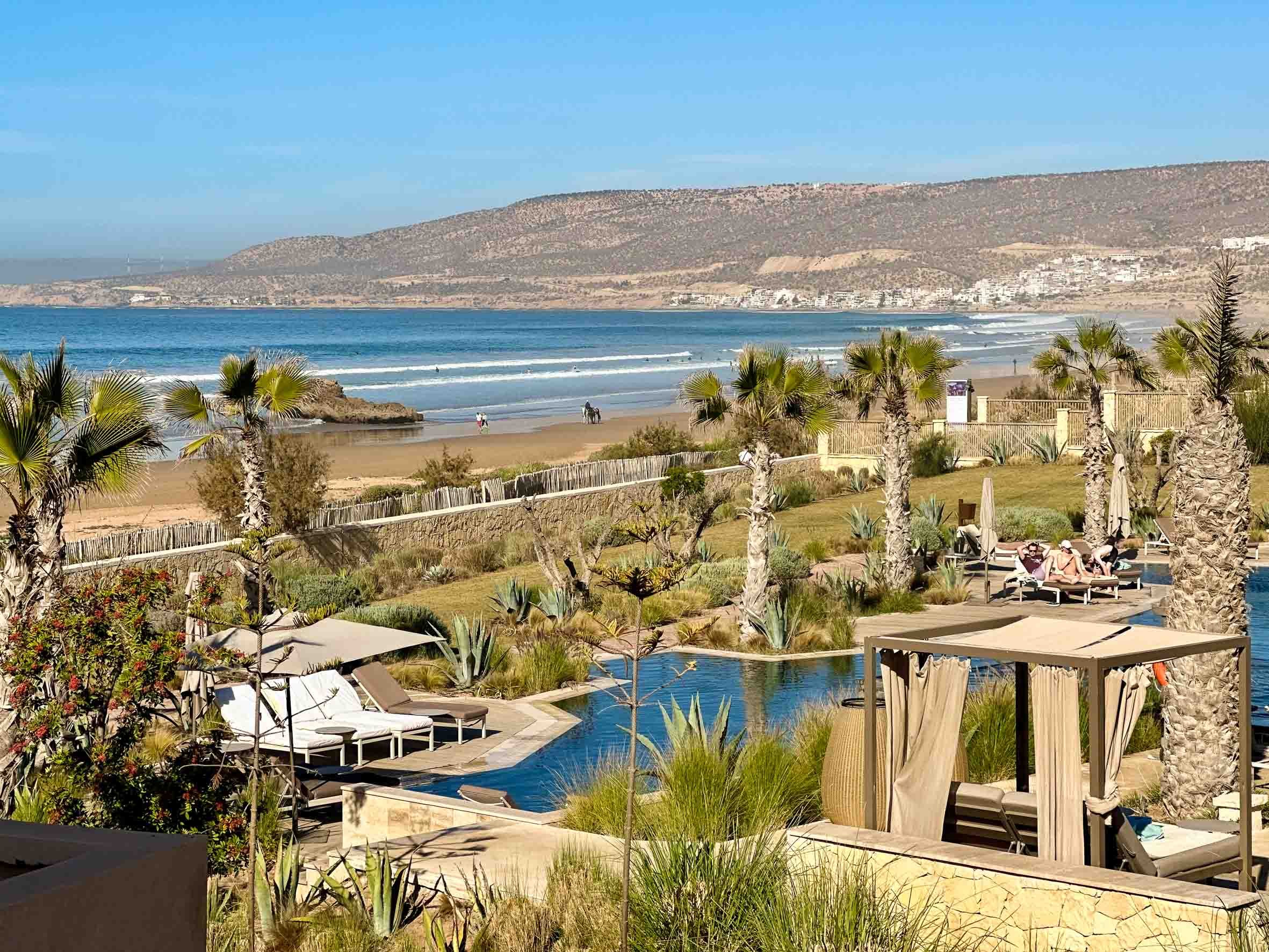 Resort pool area with palm trees, lounge chairs, and cabanas, overlooking a sandy beach with people walking by the ocean and a rocky coastline in the background.