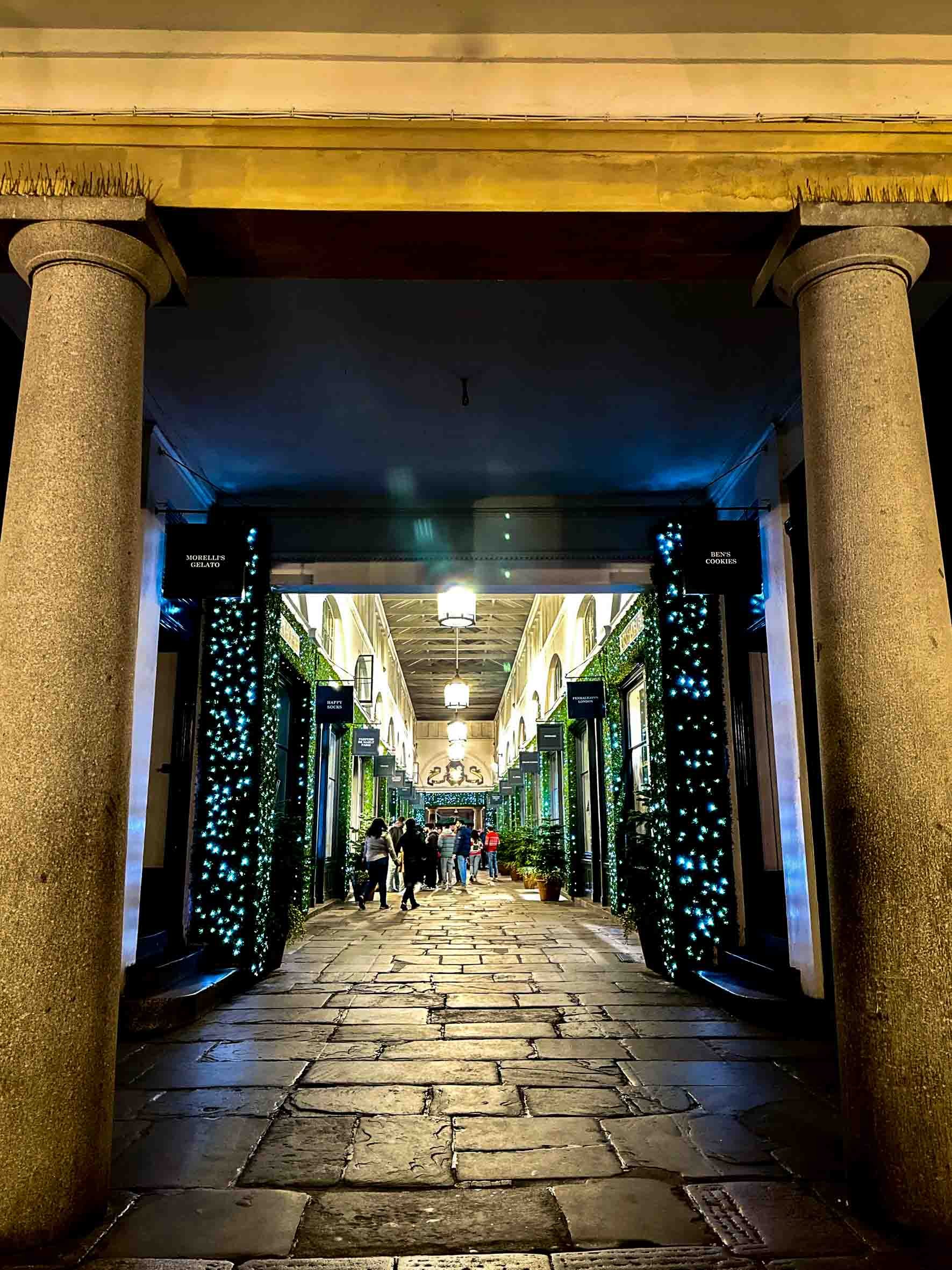 A covered outdoor shopping or dining corridor decorated with string lights, illuminated signs, and potted plants, with people walking and socializing.