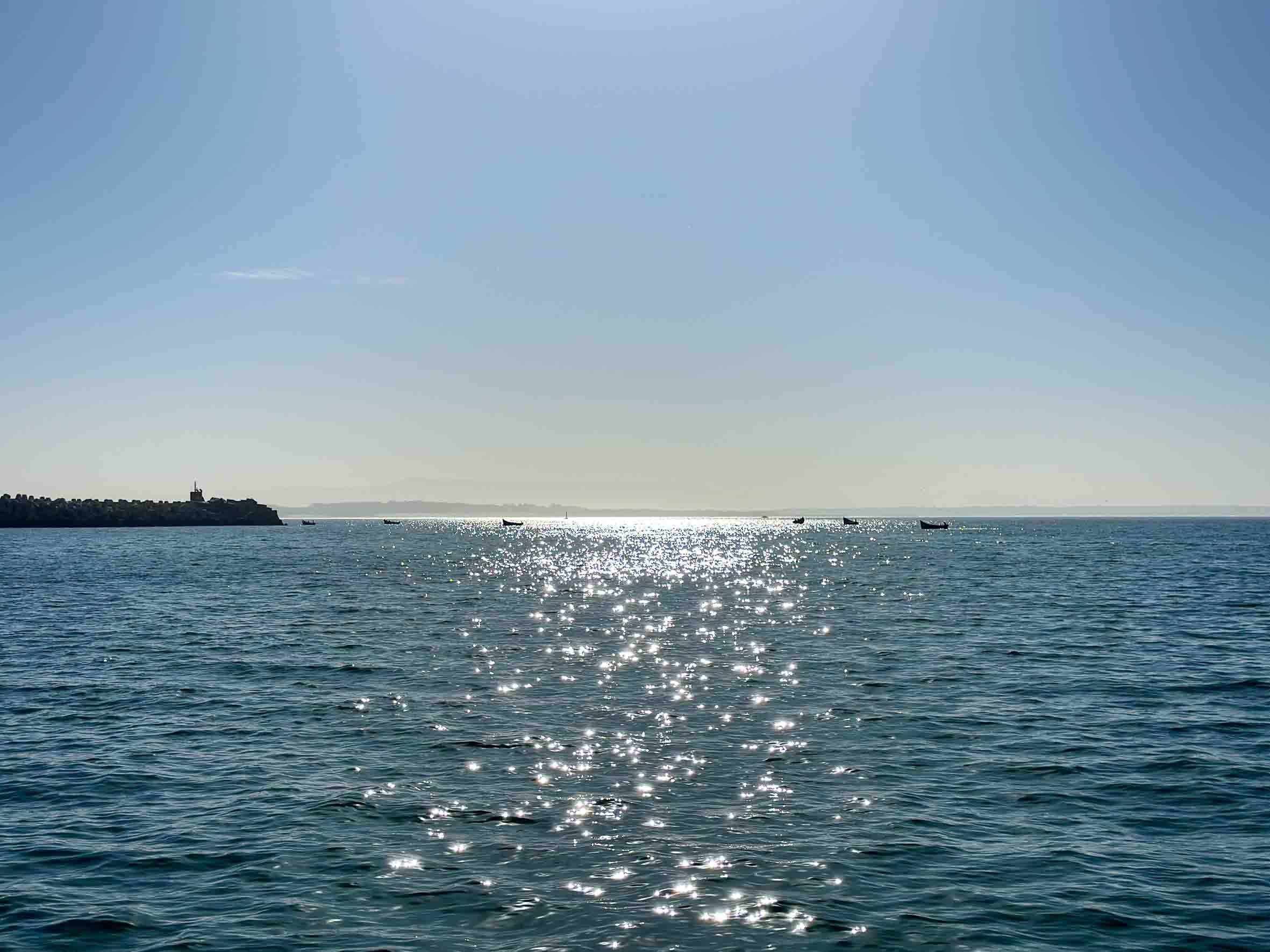 A calm ocean with gentle waves and several small boats in the distance under a clear blue sky, with the sunlight reflecting off the water.