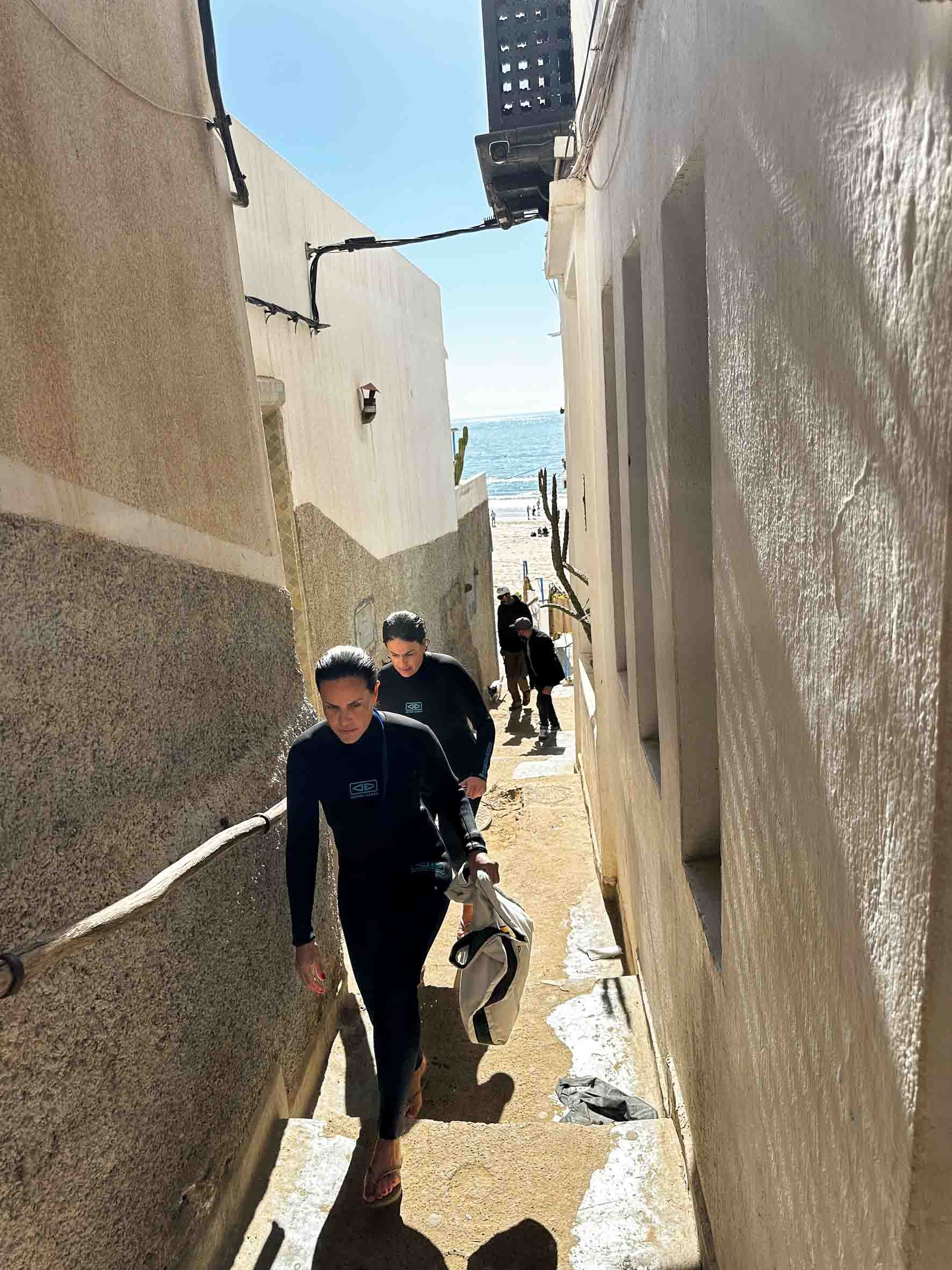 People walking up a narrow alley near the beach with the ocean visible in the background.