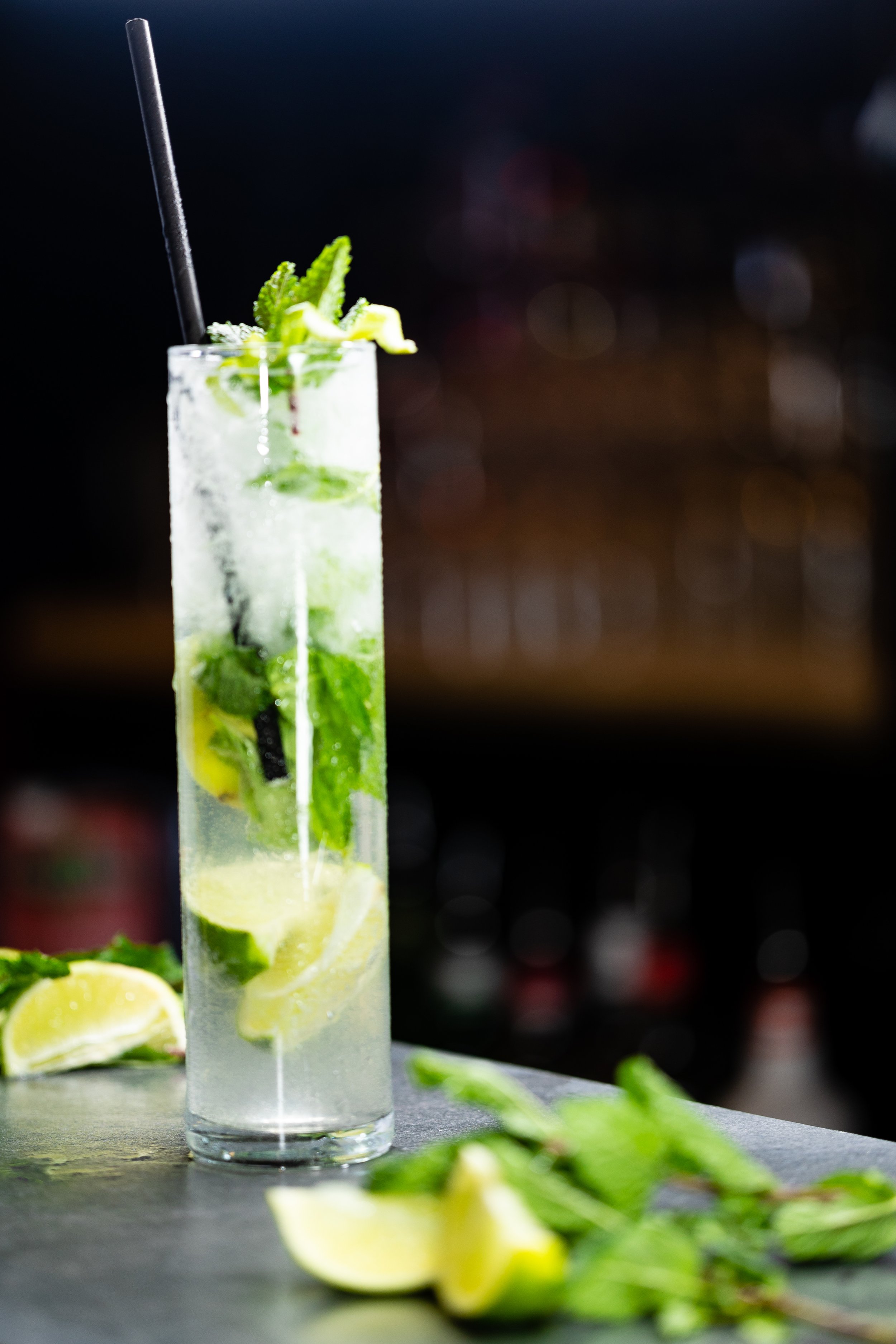 Tall glass of mojito cocktail with lime wedges, mint leaves, ice, and a black straw on a table, with lime wedges and mint scattered around.