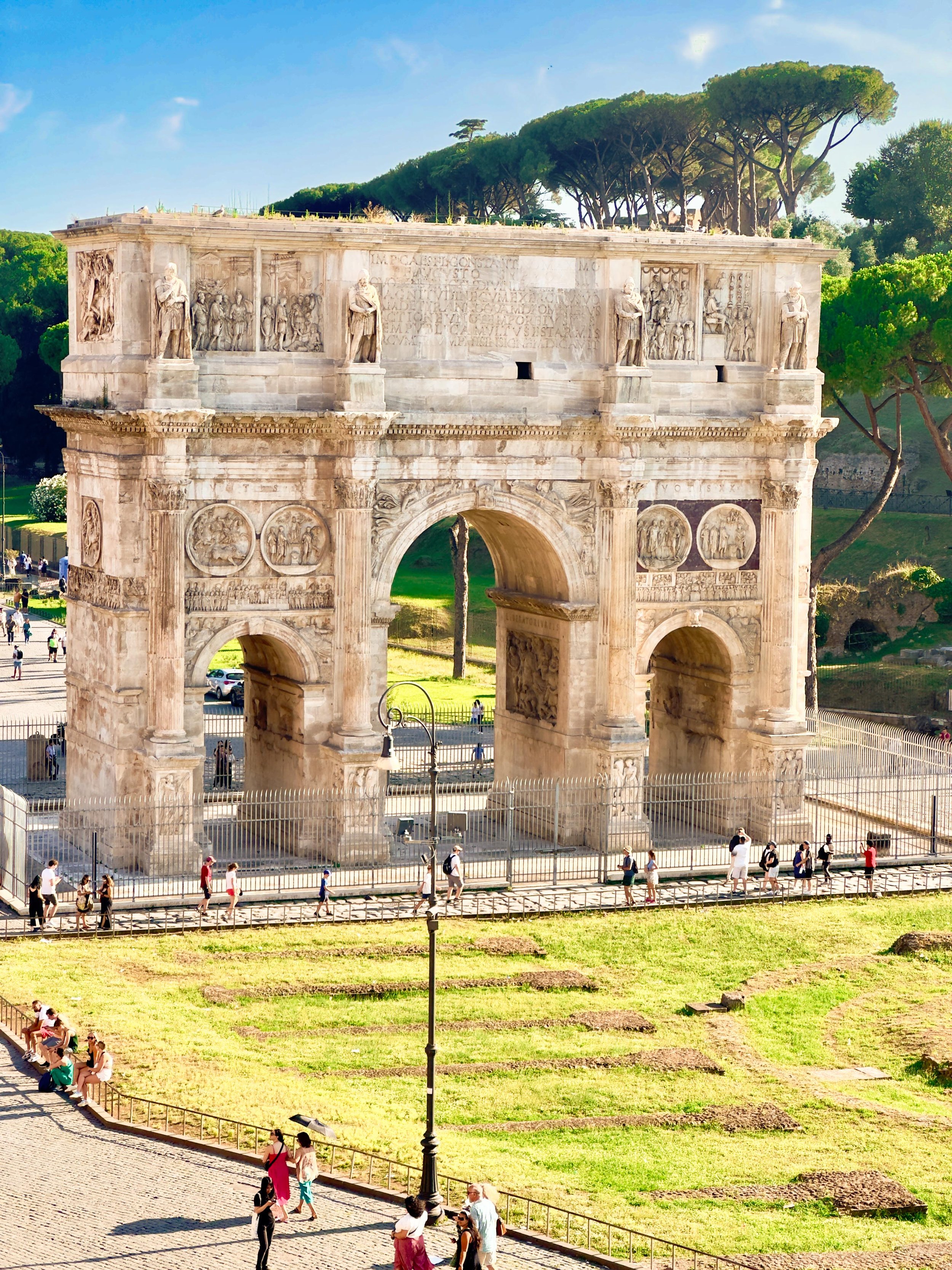 Ancient Roman triumphal arch with detailed sculptures and reliefs, surrounded by tourists and greenery.