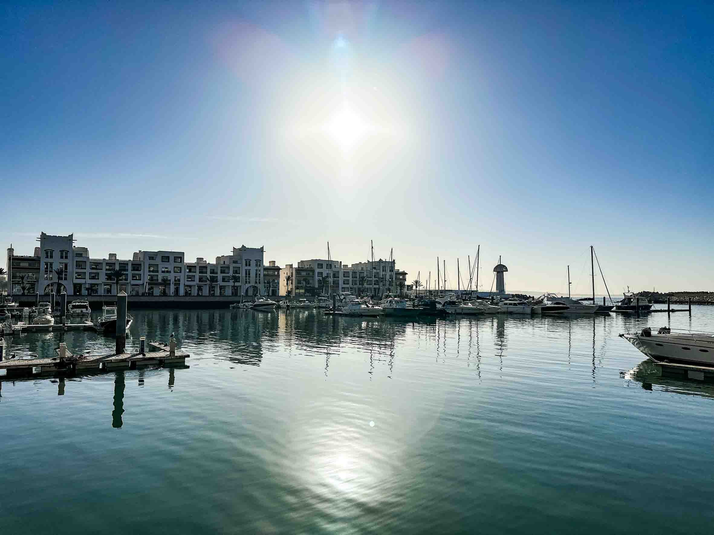 A serene marina with several boats docked, modern white buildings in the background, calm water reflecting the clear blue sky, and a lighthouse in the distance.