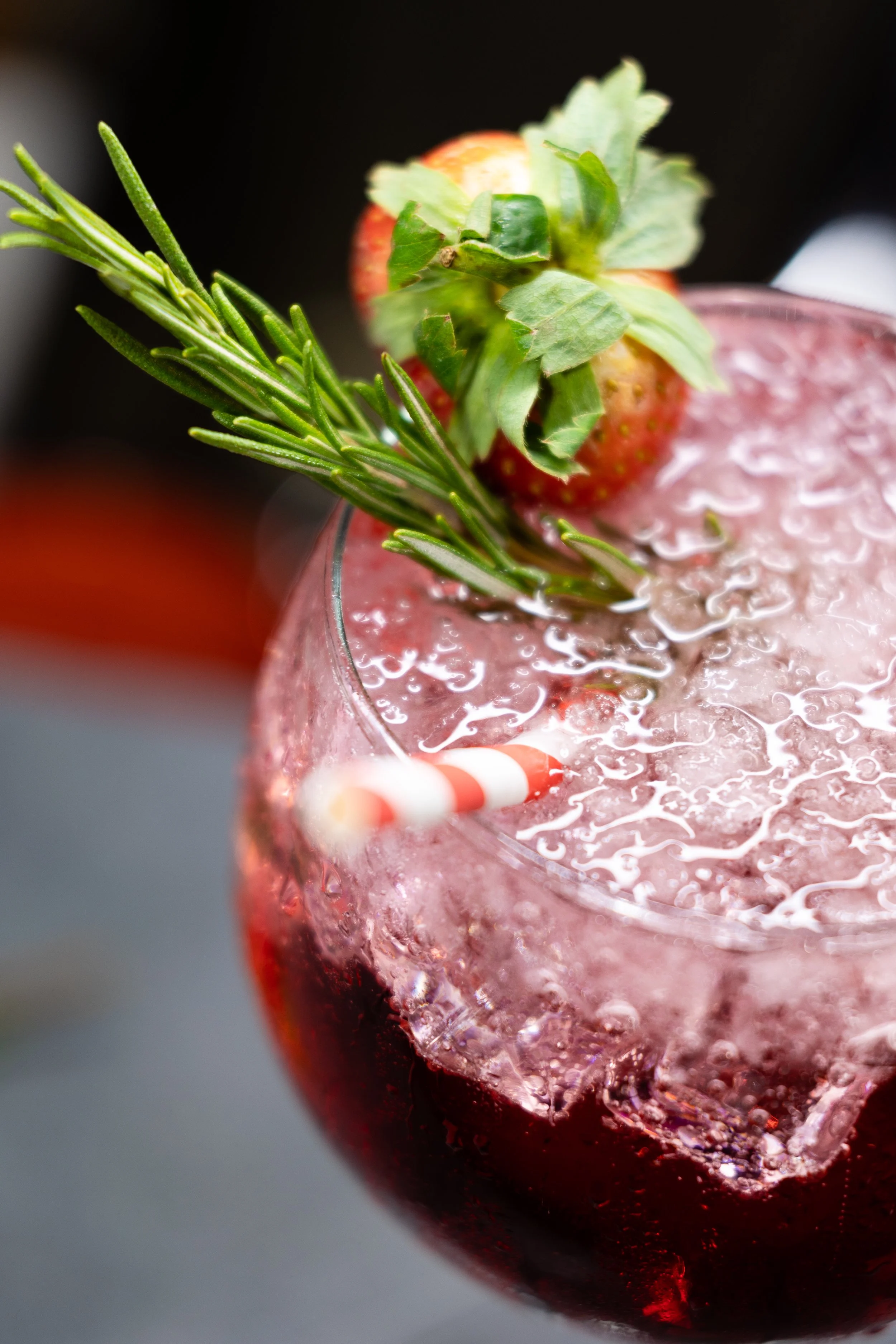 Close-up of a pink cocktail garnished with a strawberry, a sprig of rosemary, and a striped straw.