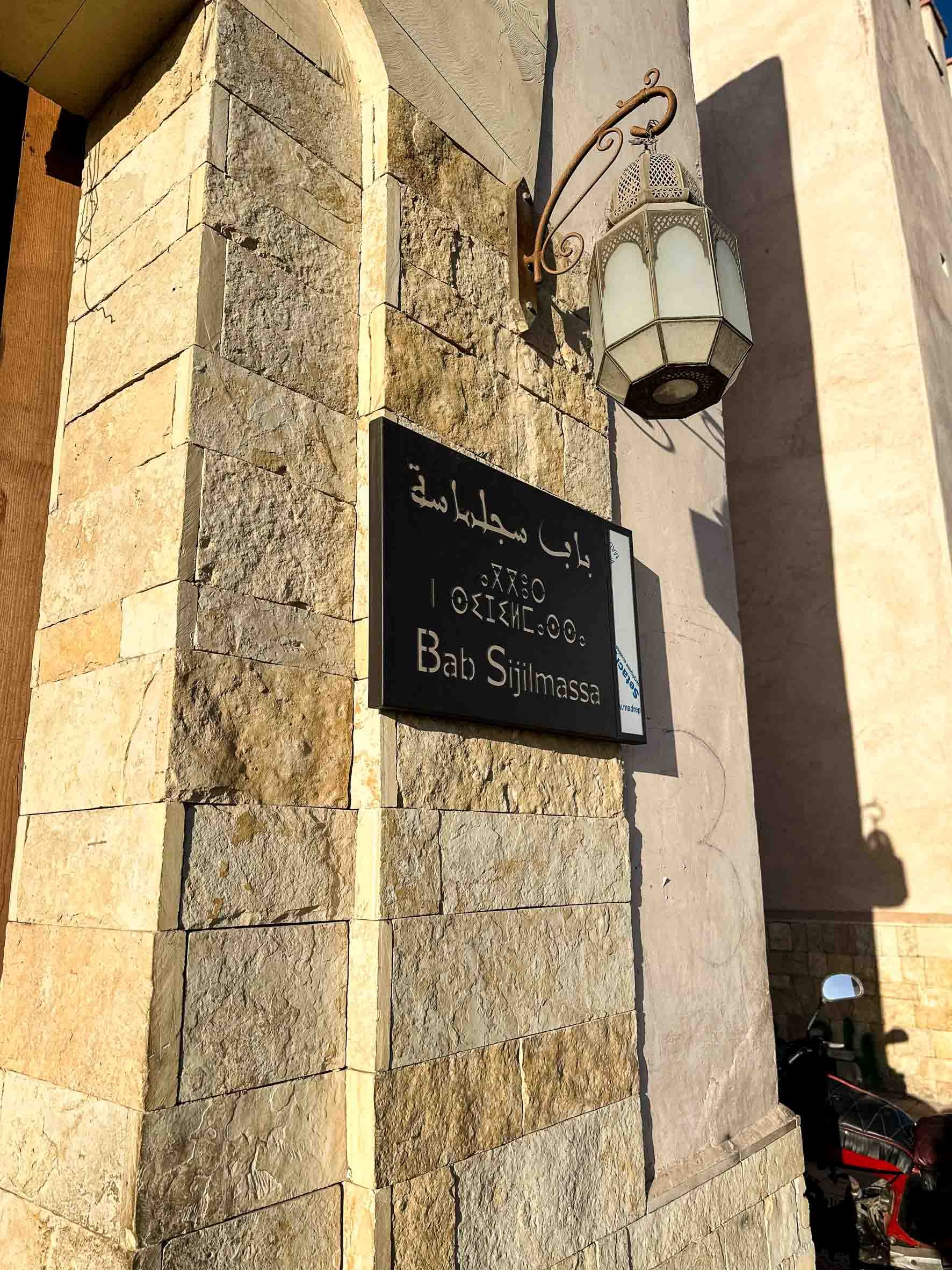 A street sign on a stone building wall displaying the name 'Bab Sijilmassa' in Arabic, English, and another language, with a decorative wall-mounted lantern above.