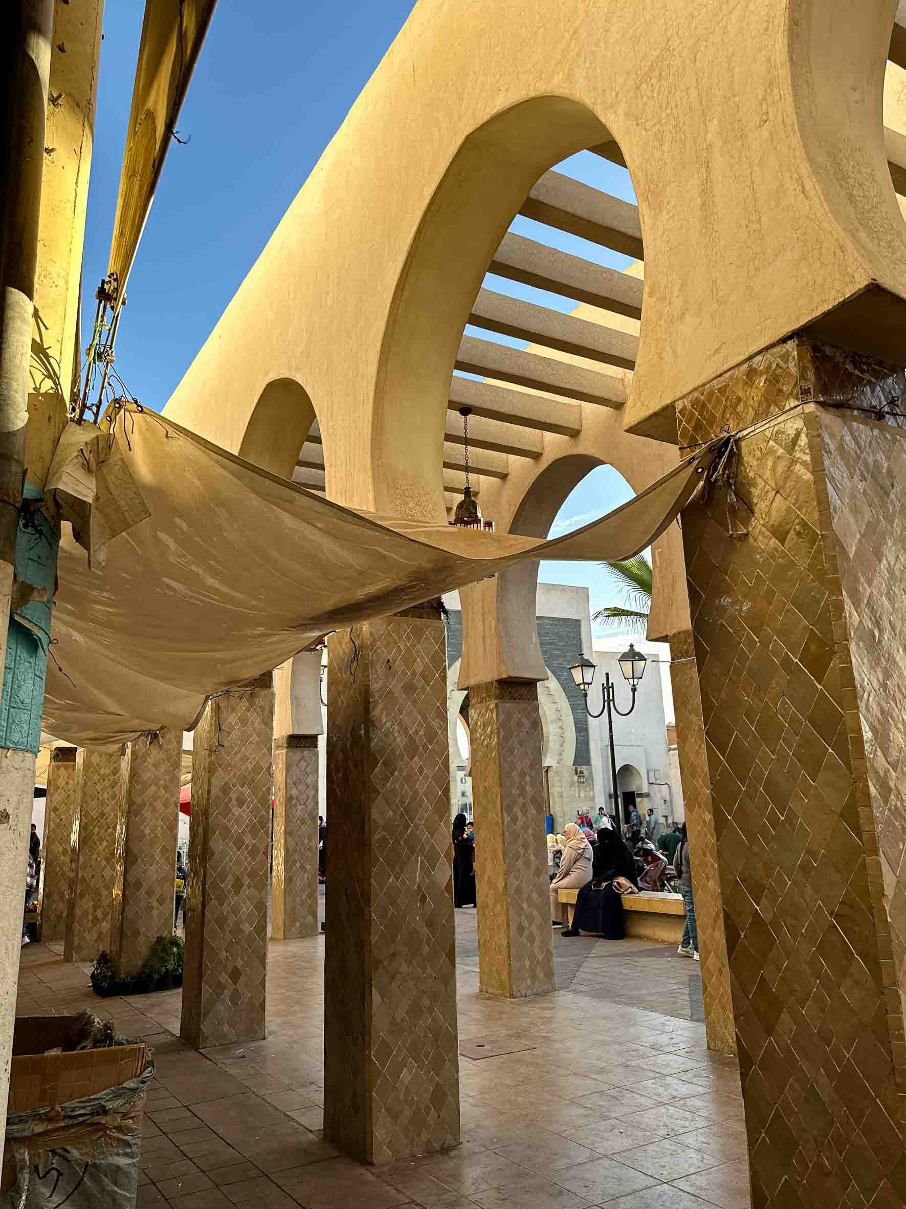 Architectural structure with large arches, golden-colored columns, and a beige fabric shade in a public area with people sitting and walking.