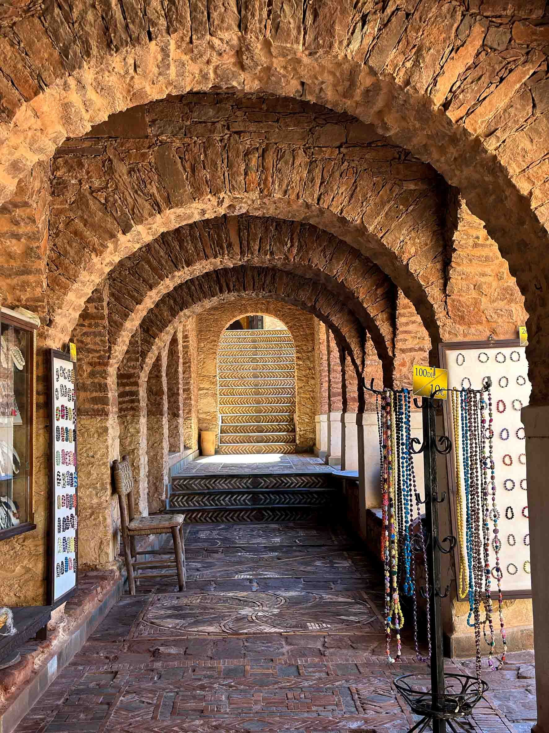 A series of brick arches in a corridor with a wooden chair, jewelry display racks with necklaces, and stairs at the end.