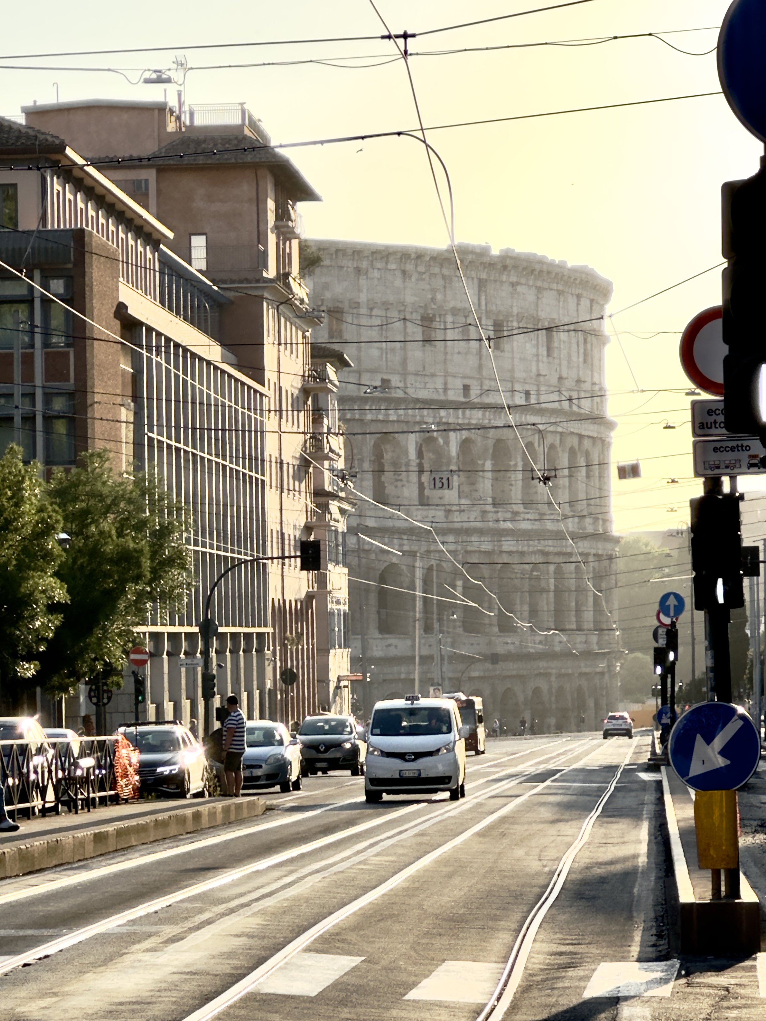 Street view with cars, buildings, and the Colosseum in the background, visible through haze or fog.