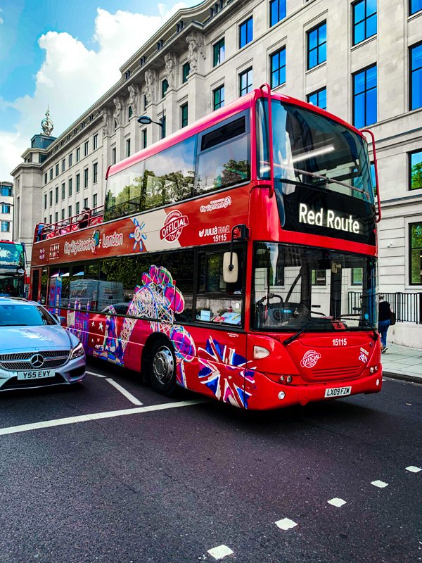 Double-decker red tour bus with 2023 London theme, displaying 'Red Route' on front, parked on city street with a silver Mercedes-Benz nearby, in front of historic white building with multiple windows.