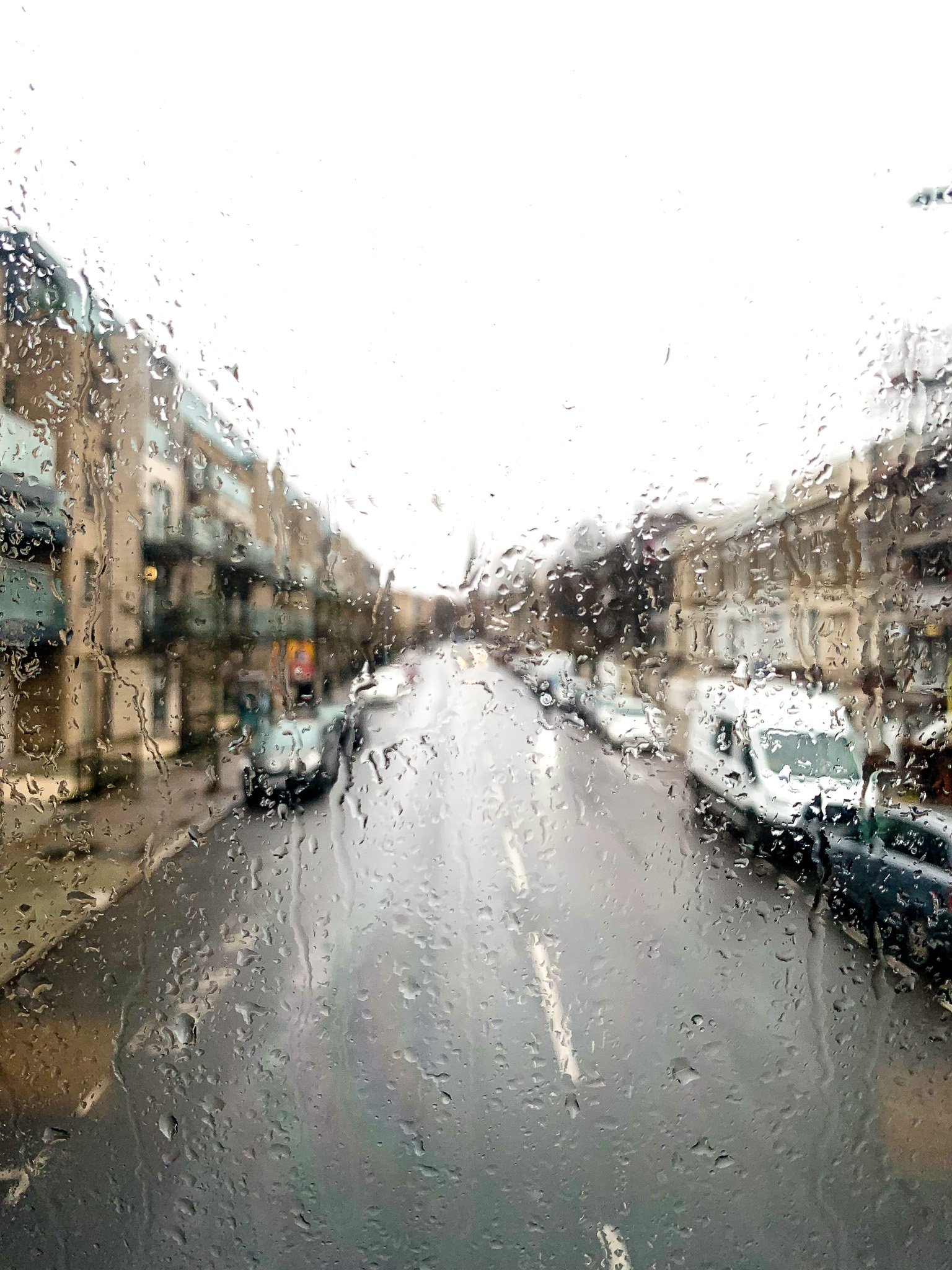 Rainy street viewed through a wet window, with parked cars and apartment buildings on both sides.