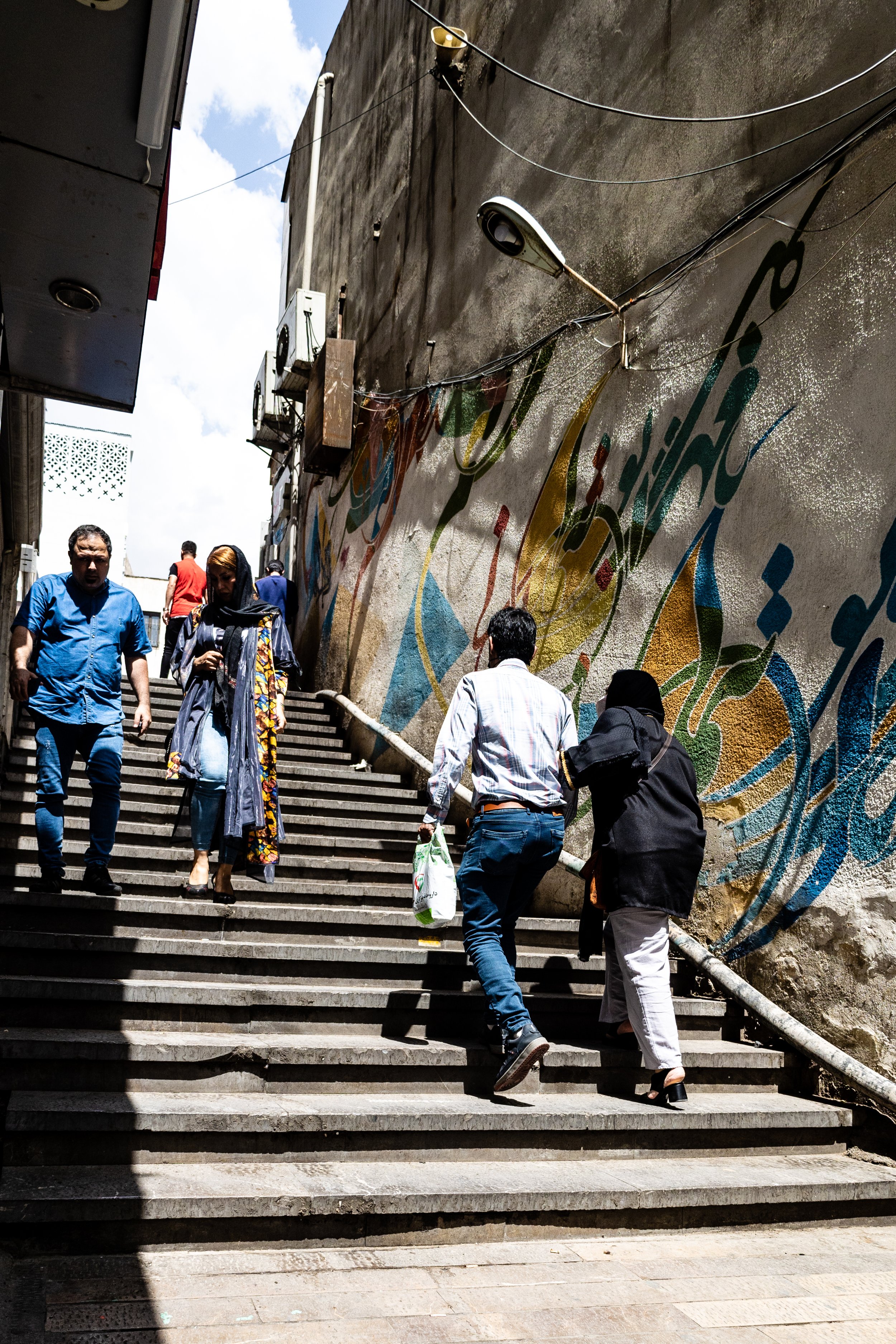 People walking up outdoor staircase beside a colorful wall mural with wires and air conditioning units.