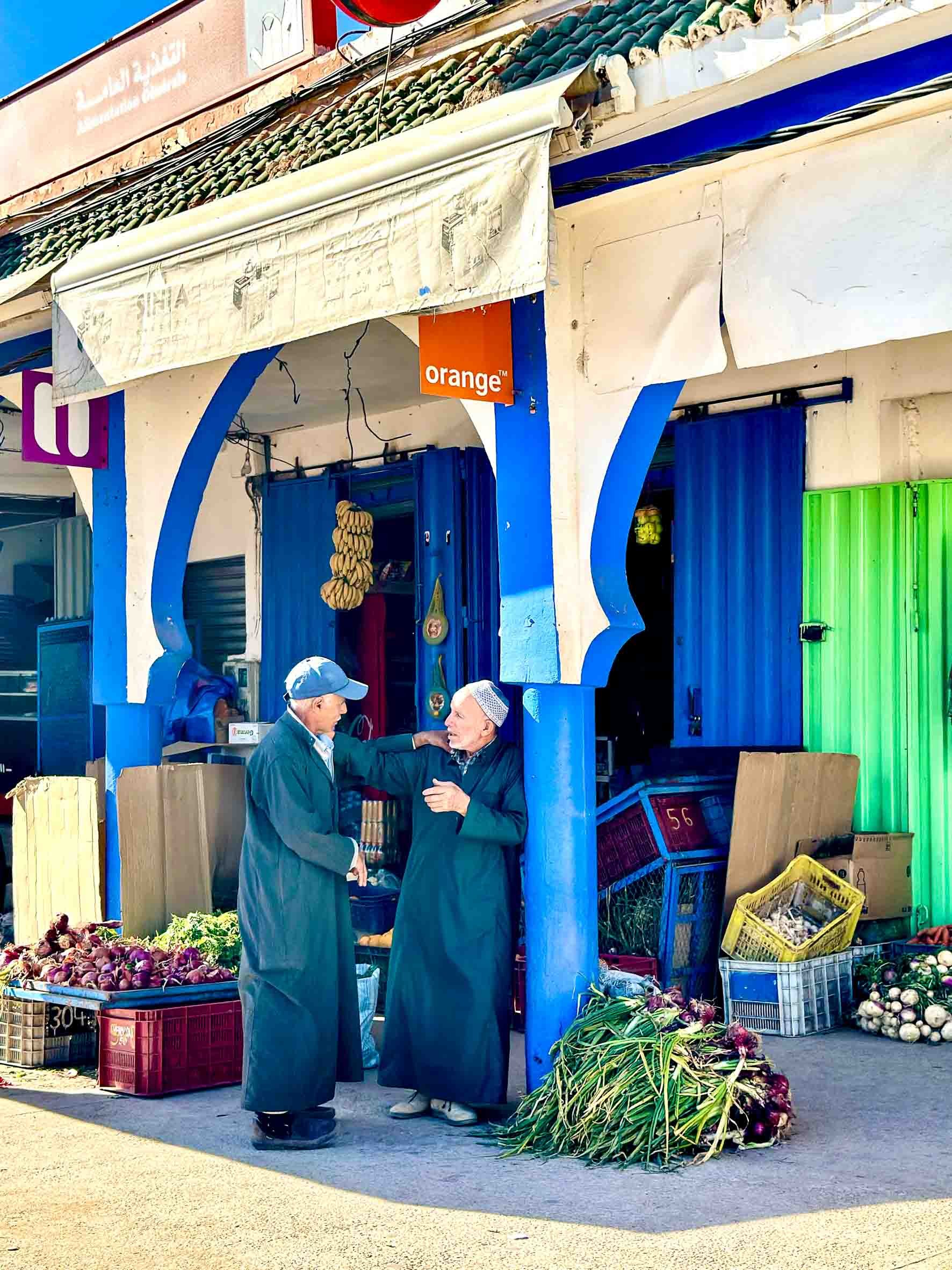 Two men in traditional robes and caps engaged in conversation outside a market stall selling onions, with crates of vegetables and colorful curtains in the background.