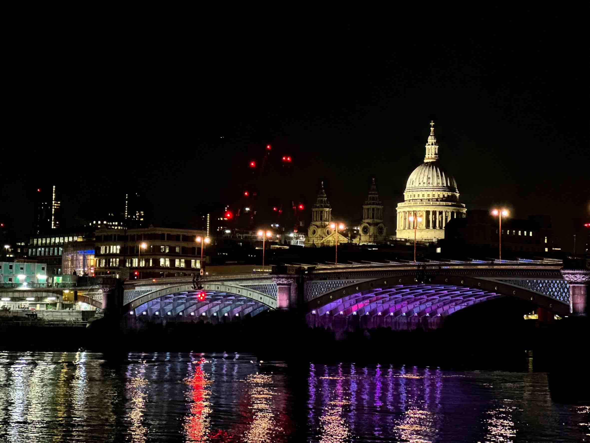 Nighttime cityscape with illuminated bridge over water and historic dome building in background.
