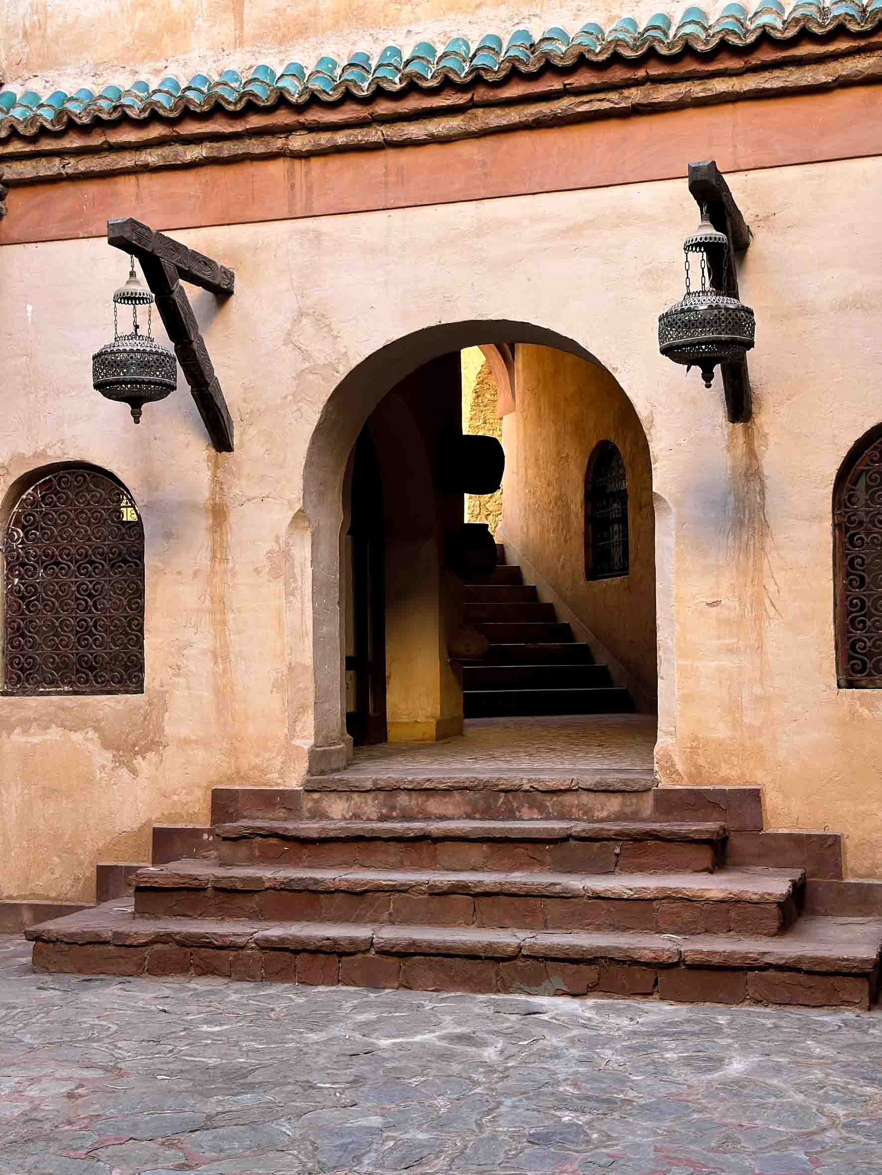 Stone steps leading up to a Mediterranean-style building entrance with an arched doorway, two small barred windows on either side, and decorative hanging lanterns.