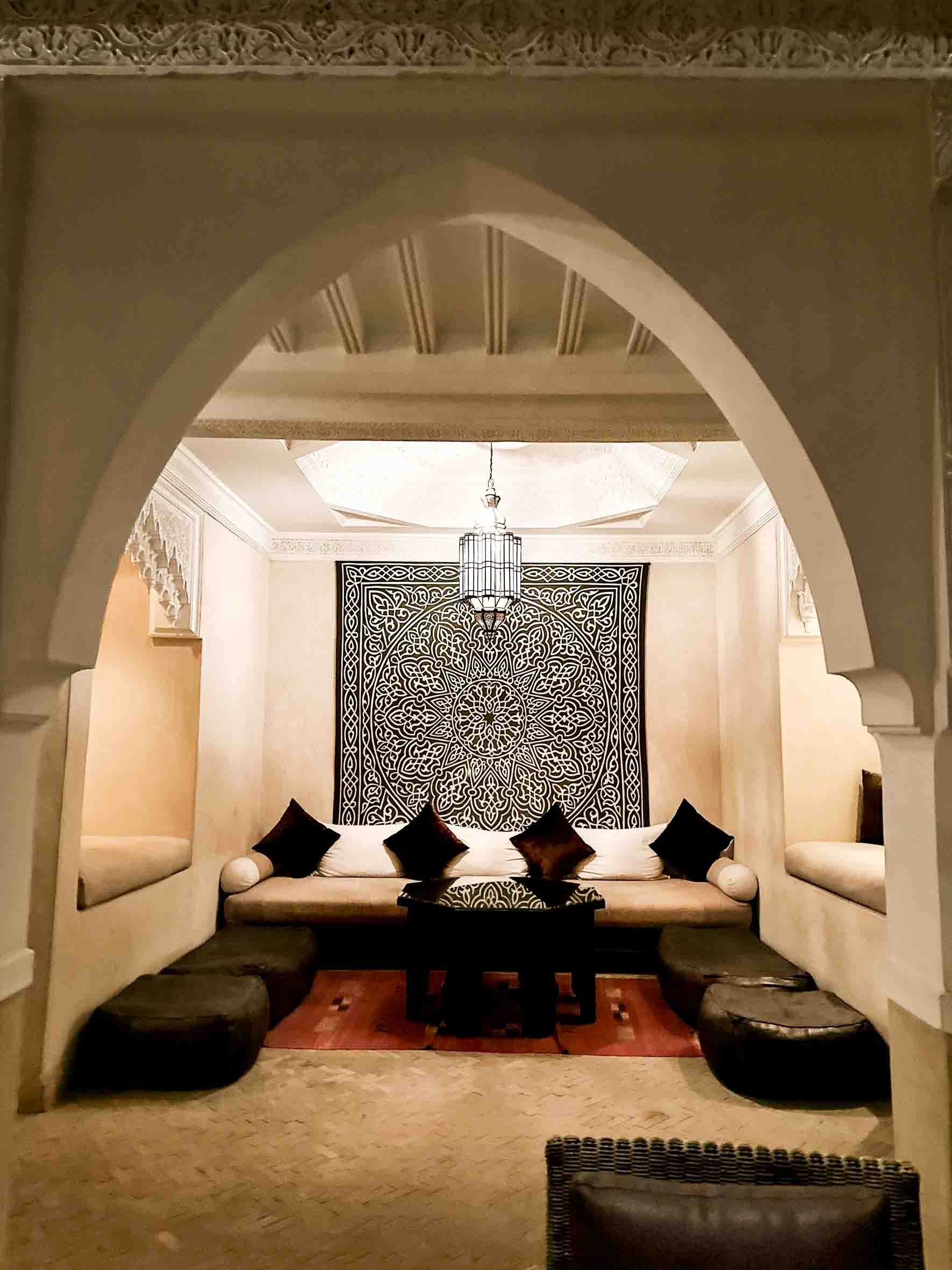 Living room with Middle Eastern decor, including a wall hanging with intricate black and white geometric pattern, a large chandelier, a white sofa with black and brown pillows, and built-in seating areas