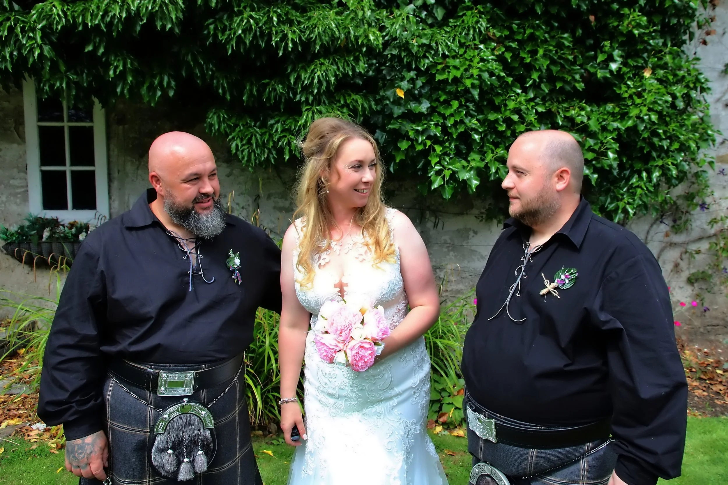 A bride in a white wedding dress holding a bouquet of pink flowers, standing between two men in black shirts wearing kilts, outdoors with green foliage and a stone wall