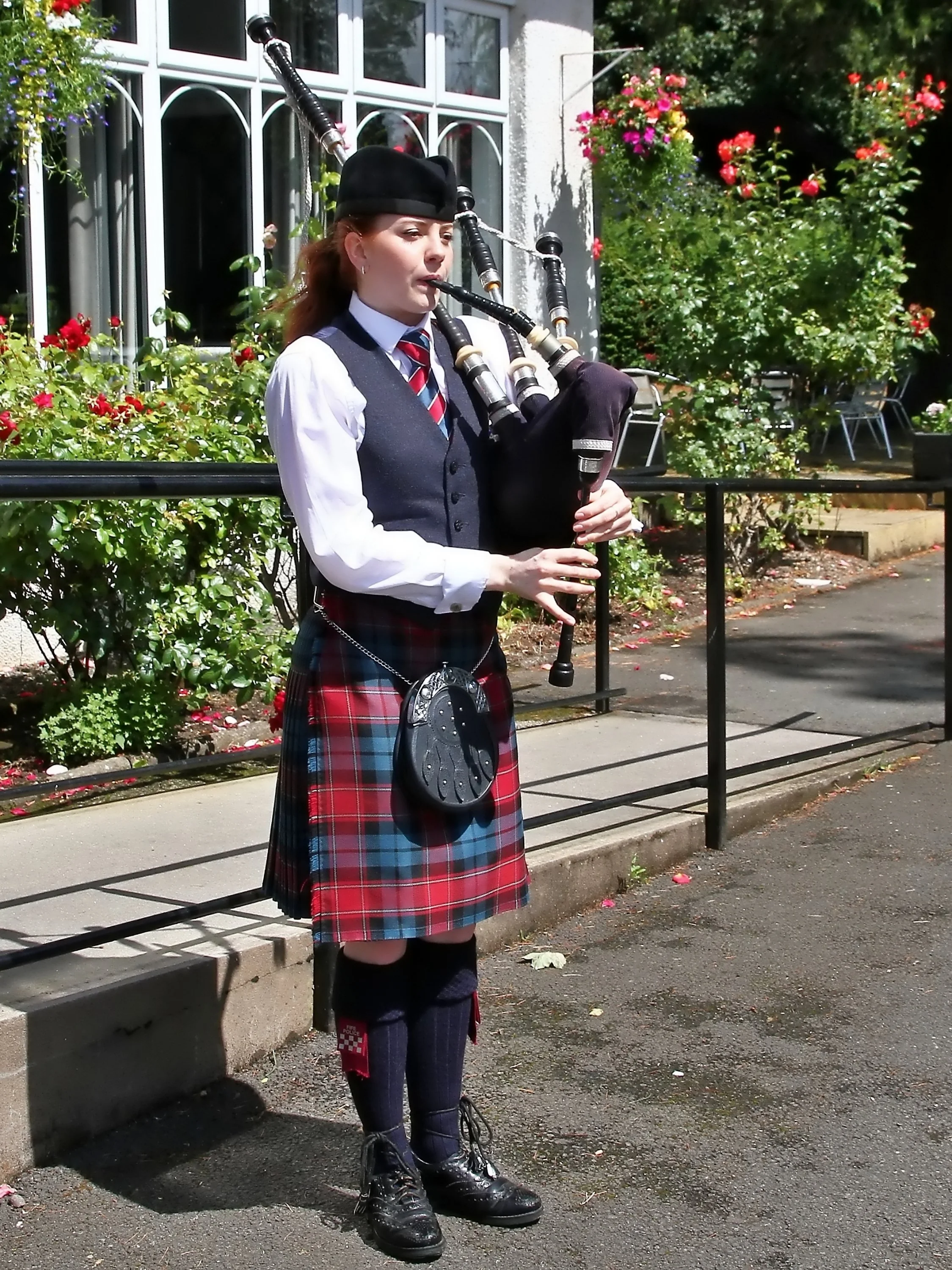 Young woman in traditional Scottish attire playing bagpipes outdoors, surrounded by flowers and buildings.