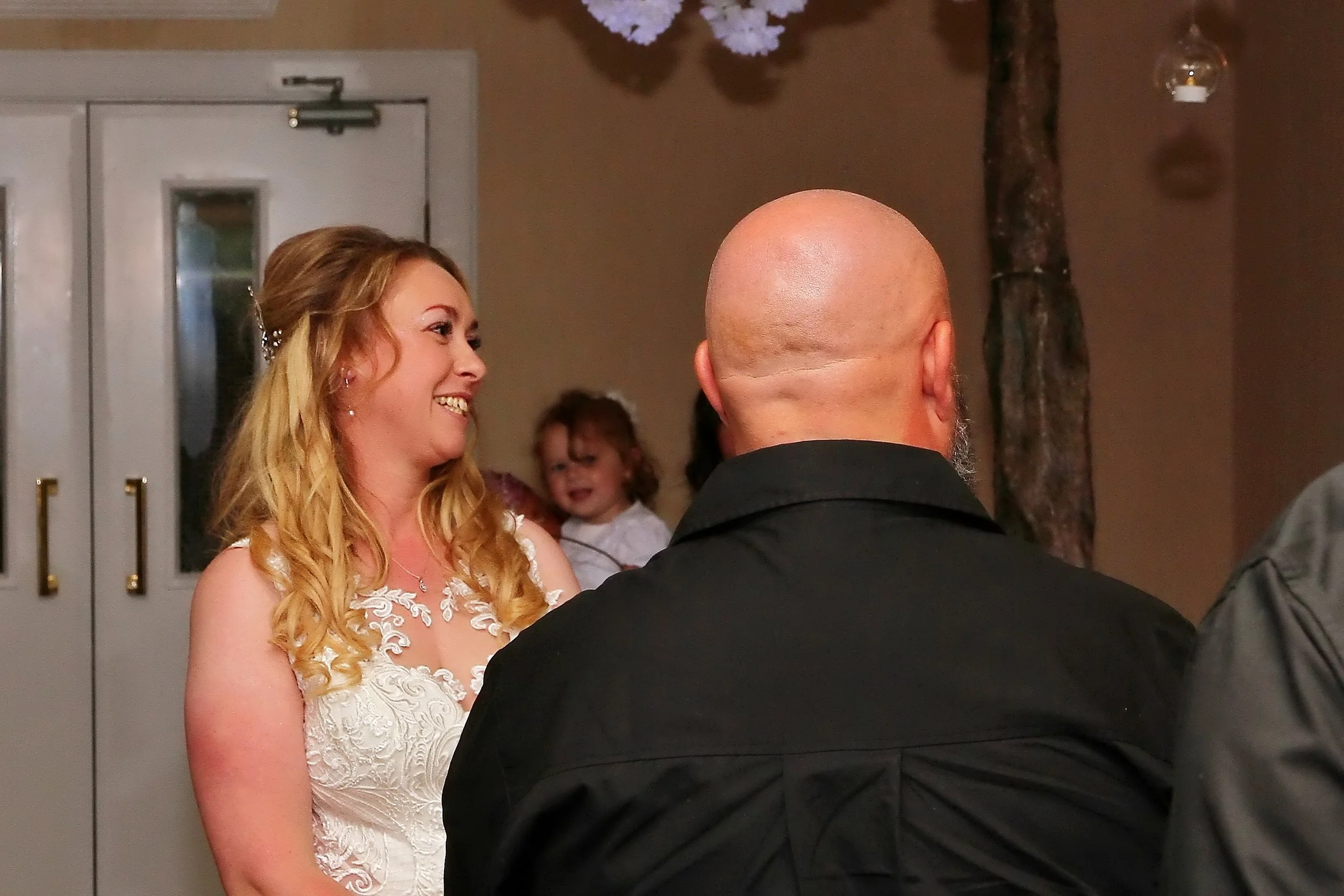 A woman in a white dress with lace details smiling and talking to a bald man in a black shirt, with a young girl in the background.