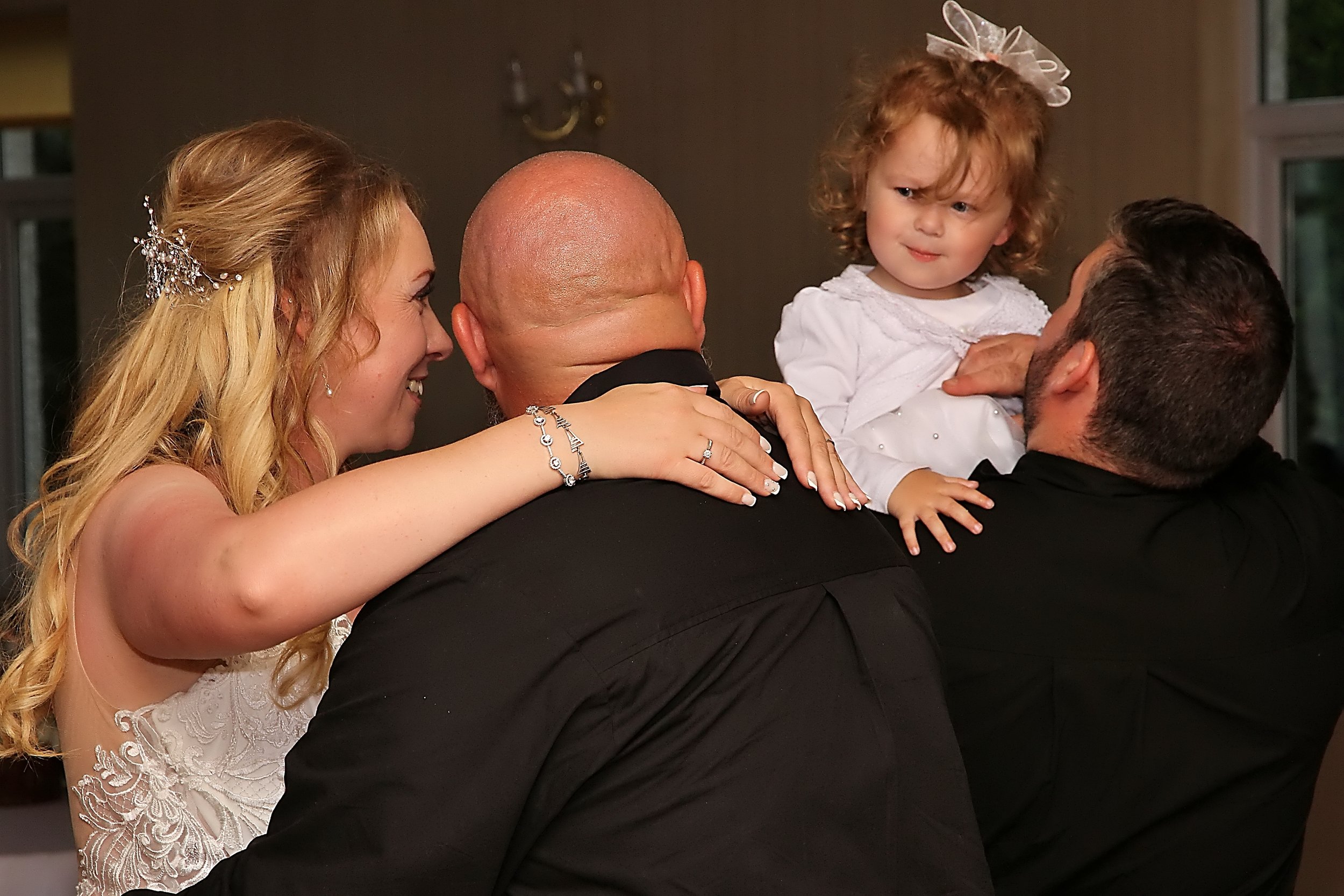 A woman and a man are holding two young girls, with the woman smiling and the girl looking confused or annoyed at the man. The girls are wearing white dresses, and the woman has long blonde hair with a decorative hairpiece. The man and woman are dres