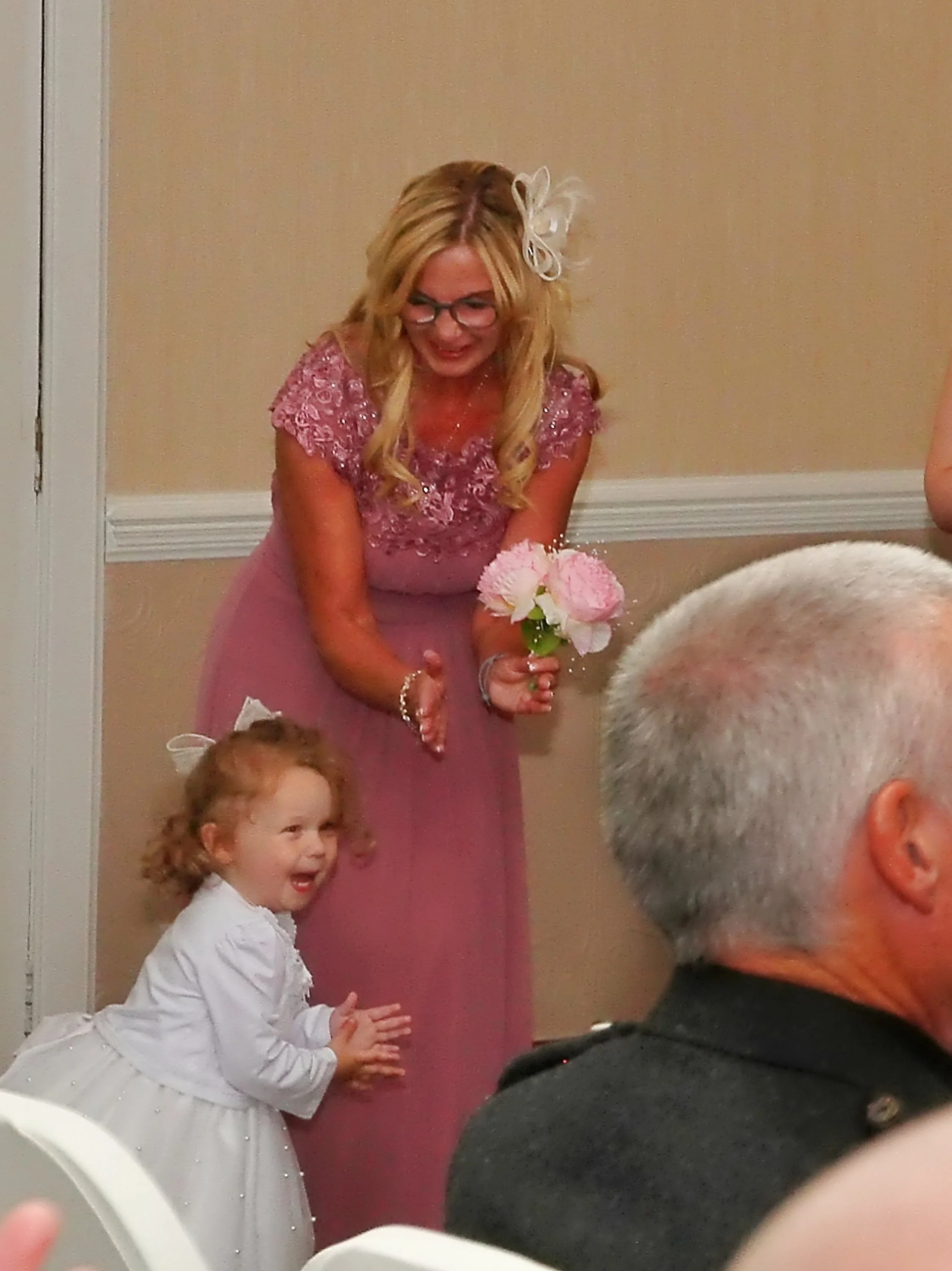 A woman in a pink dress holding pink flowers, smiling and gesturing, with a young girl in a white dress clapping and smiling beside her, at an indoor celebration or event.