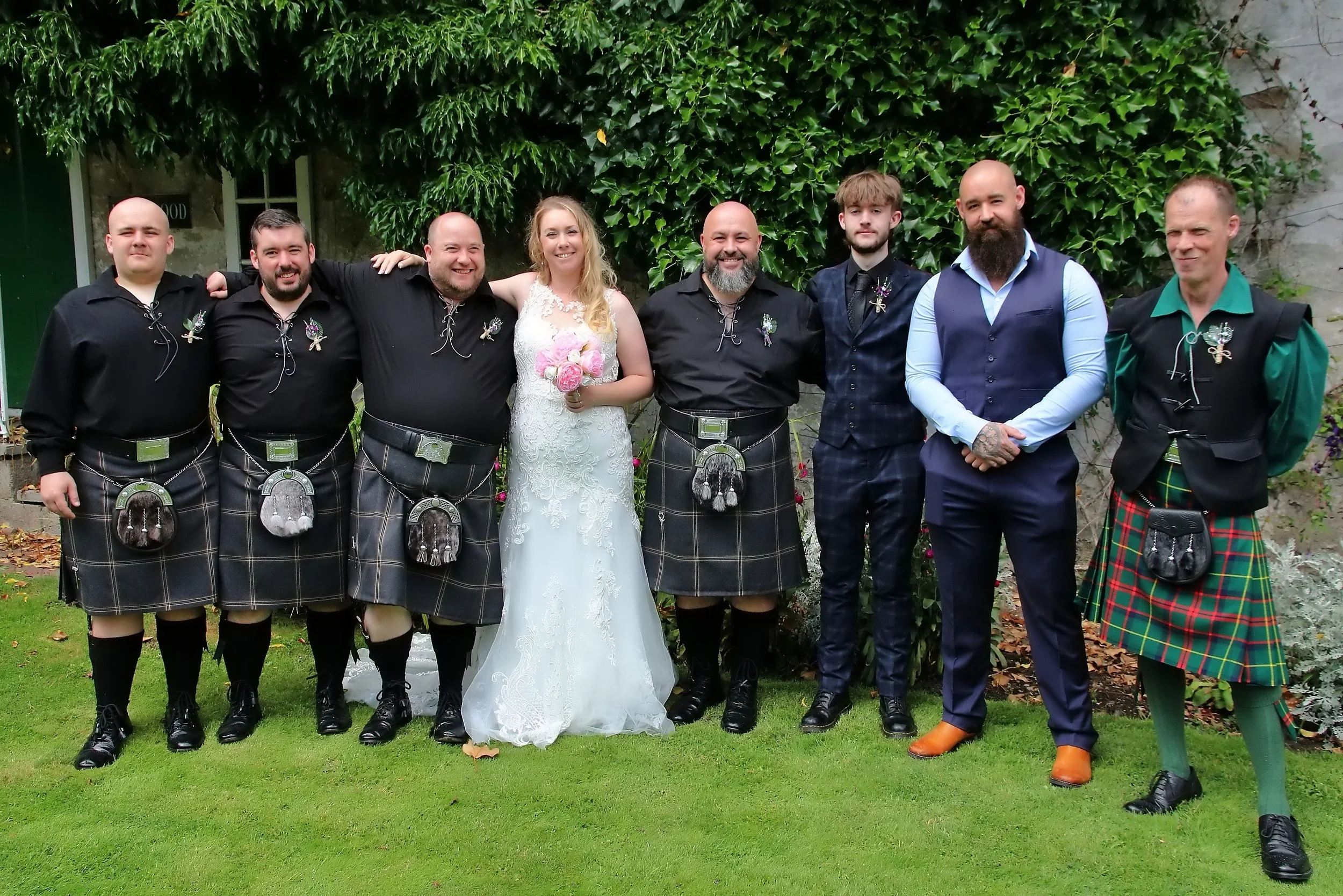 Group of people standing on grass, dressed in wedding attire with some in kilts, outdoors with green foliage background.