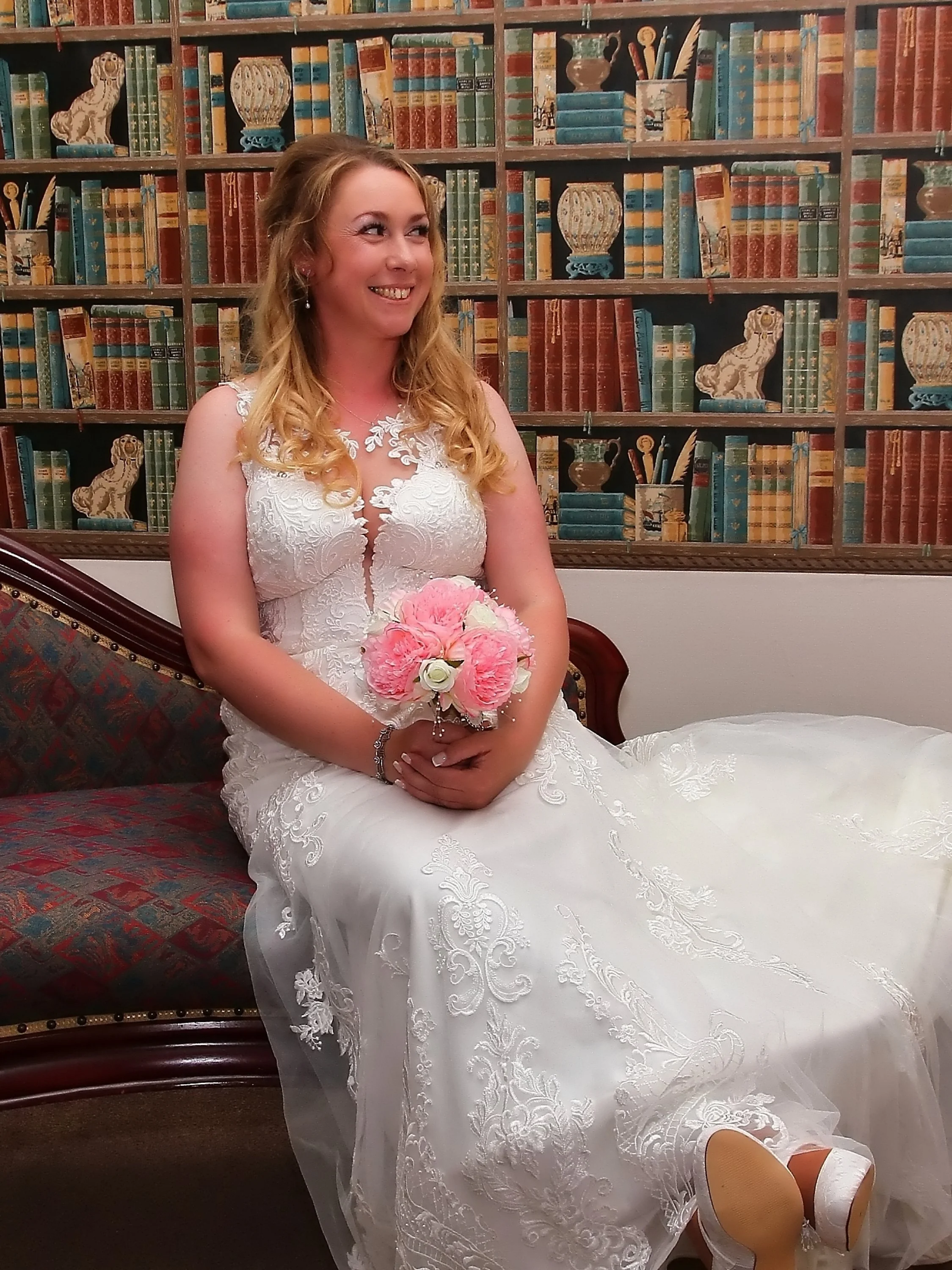 A smiling woman in a white wedding dress holding a pink bouquet, sitting on a vintage-style sofa, with a bookshelf wallpaper background.