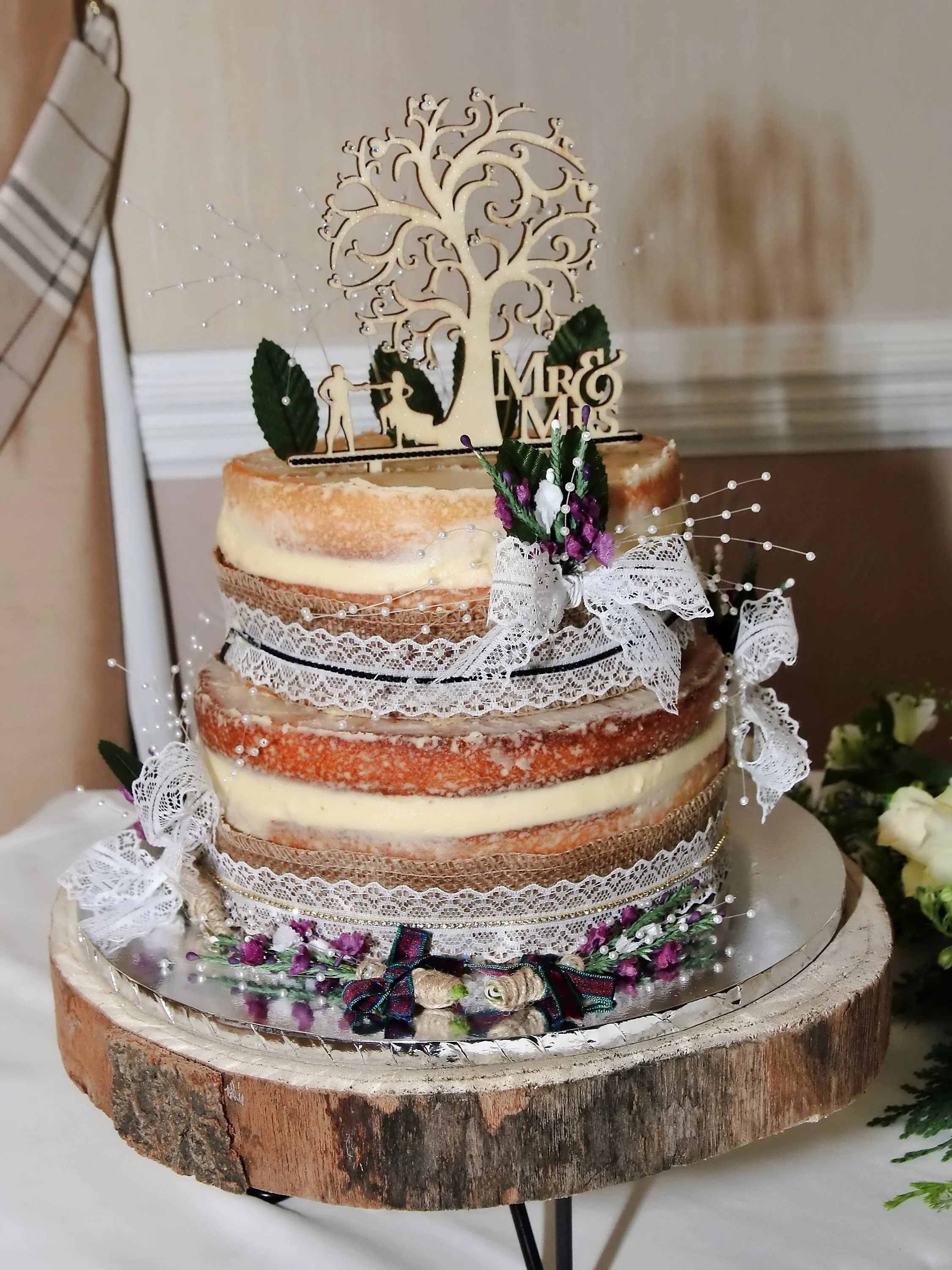 Naked wedding cake with lace and ribbon decorations, topped with a wooden couple and tree cake topper, on a wooden slice