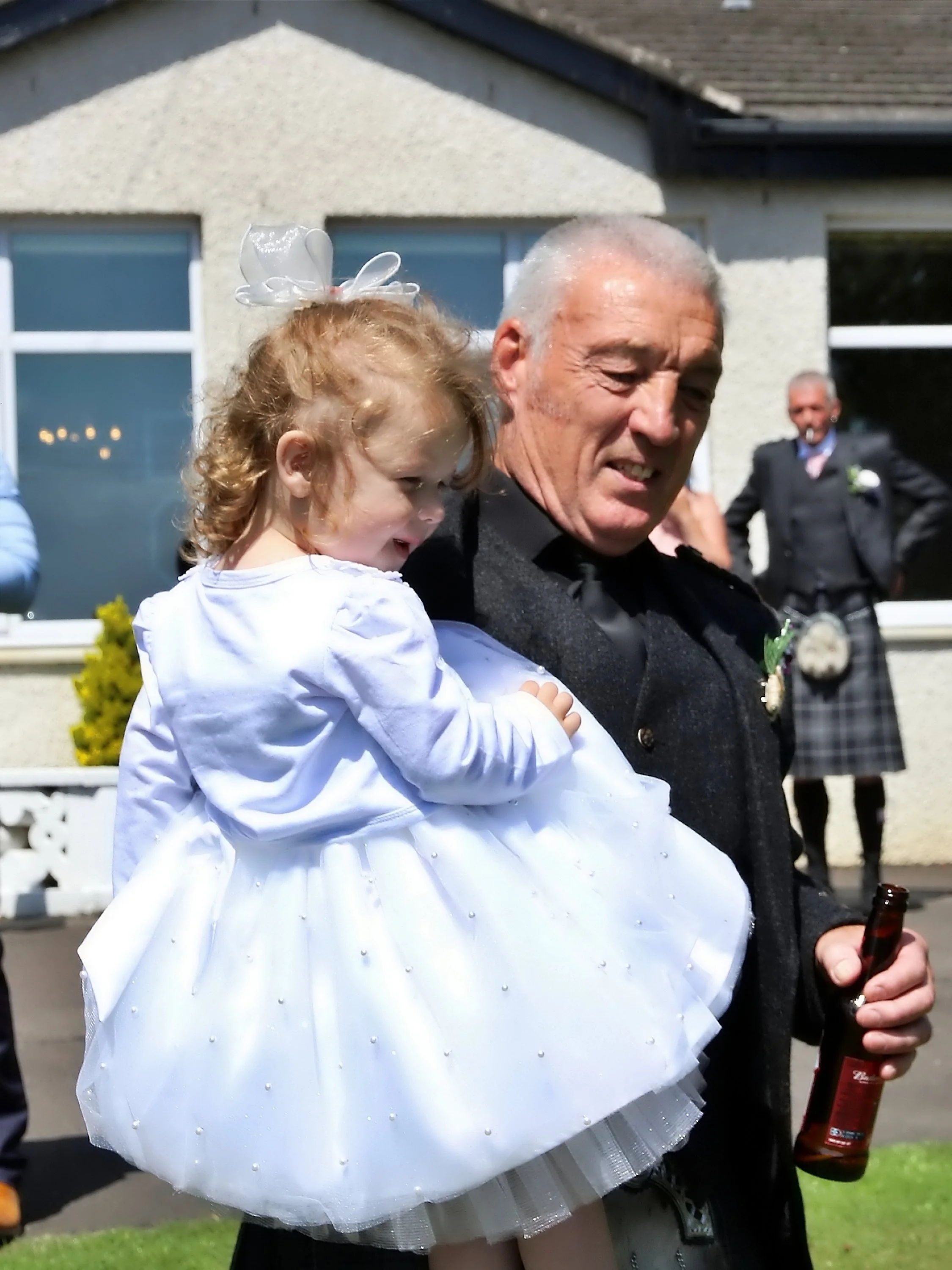 An older man holding a young girl in a white dress, outside a house on a sunny day, with other people in the background.