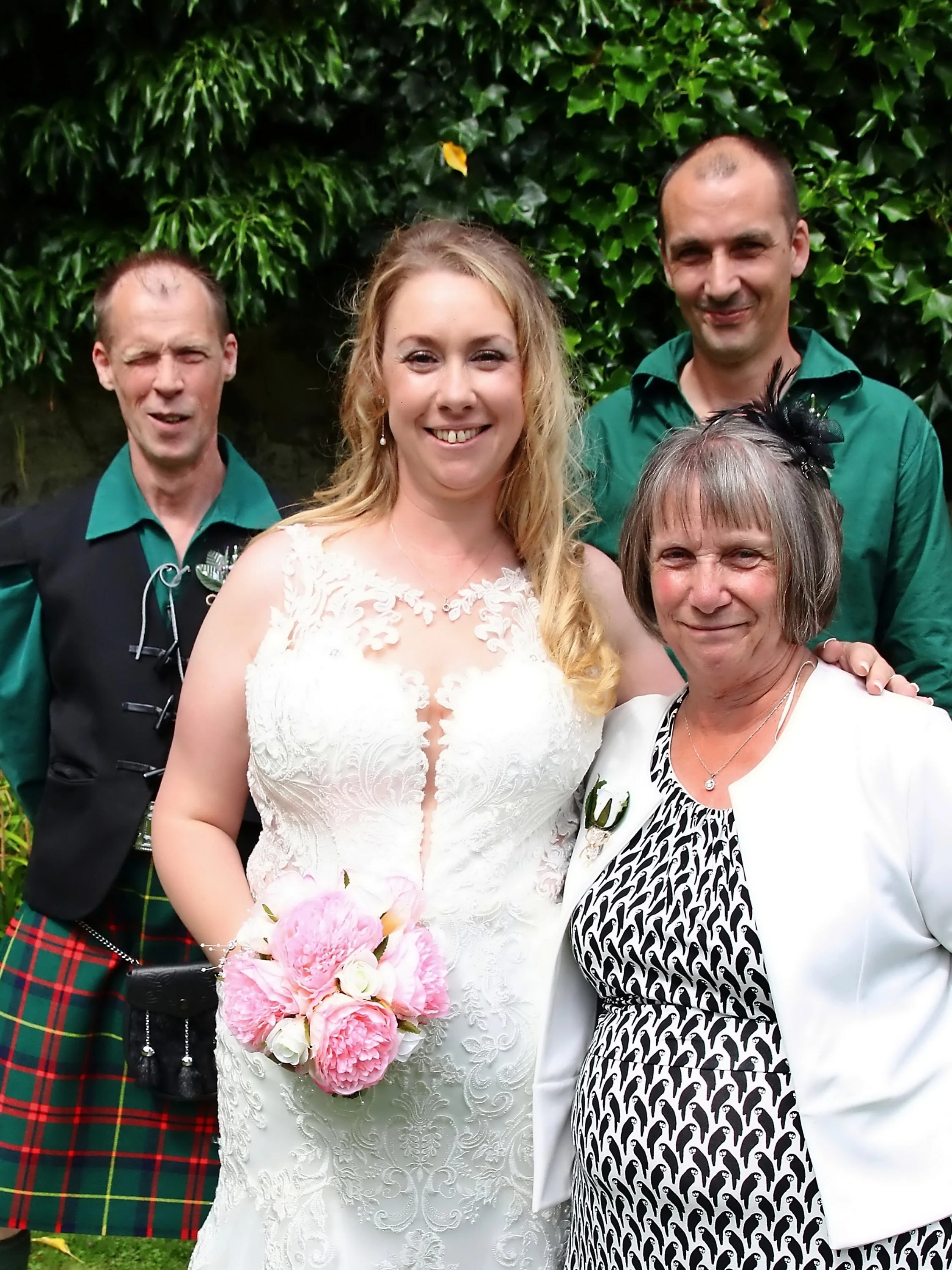 A bride in a white lace wedding dress holding a pink and white bouquet standing with four other people in front of a green leafy background at a wedding.