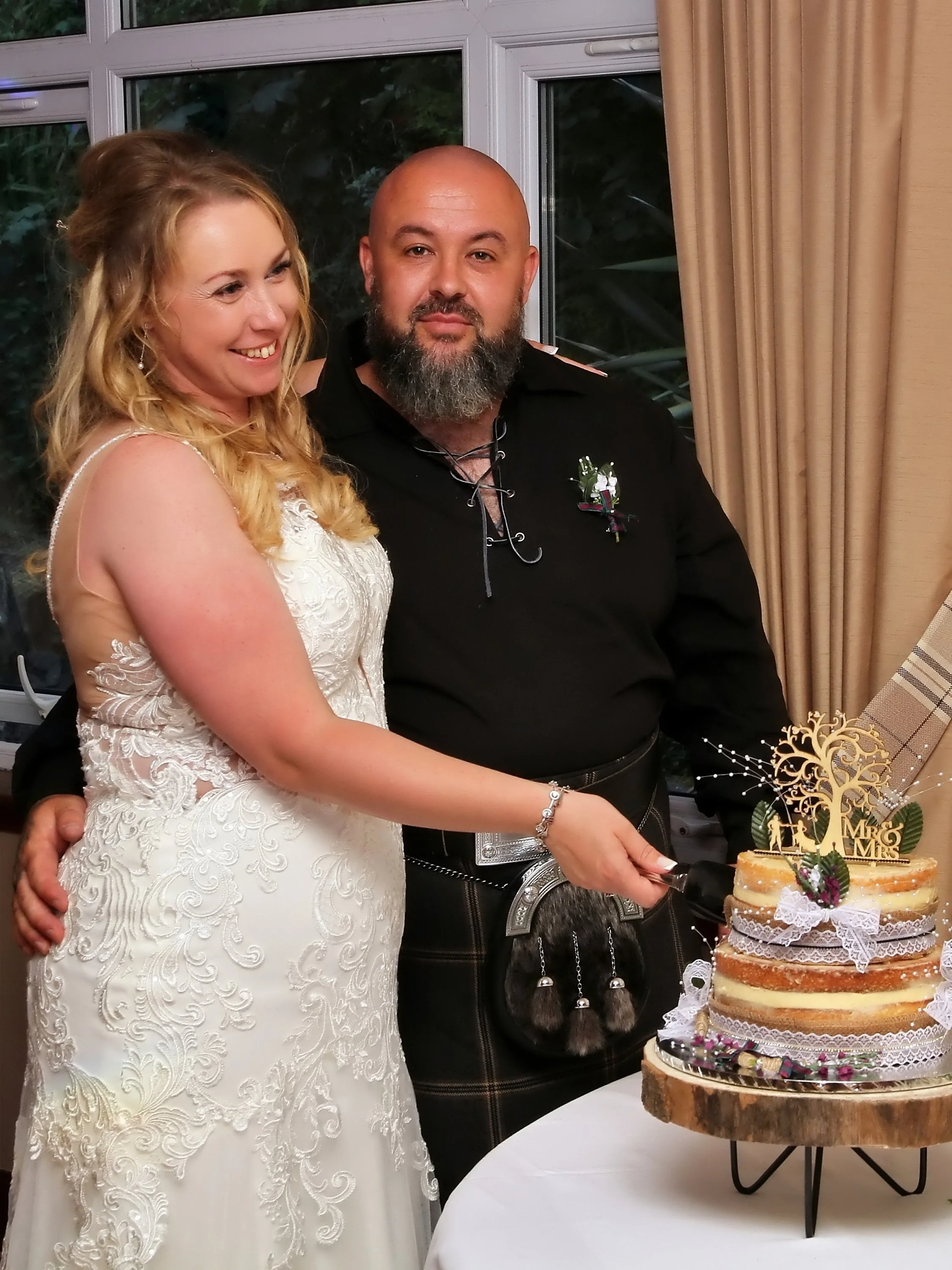 A bride and groom cutting a wedding cake together, celebrating their wedding, with the bride in a white lace gown and the groom in traditional Scottish attire, inside a decorated venue.