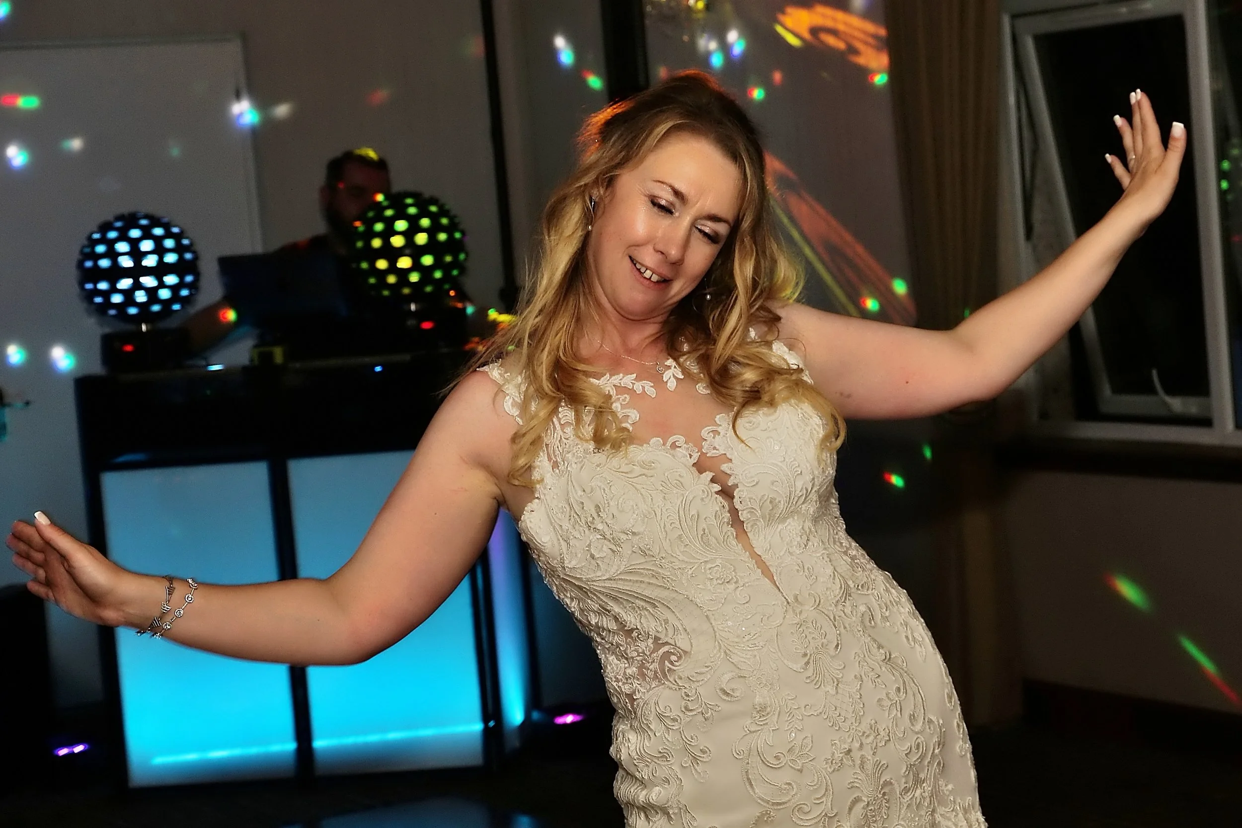 Woman dancing in a white lace dress at a party with colorful disco lights and a DJ in the background.