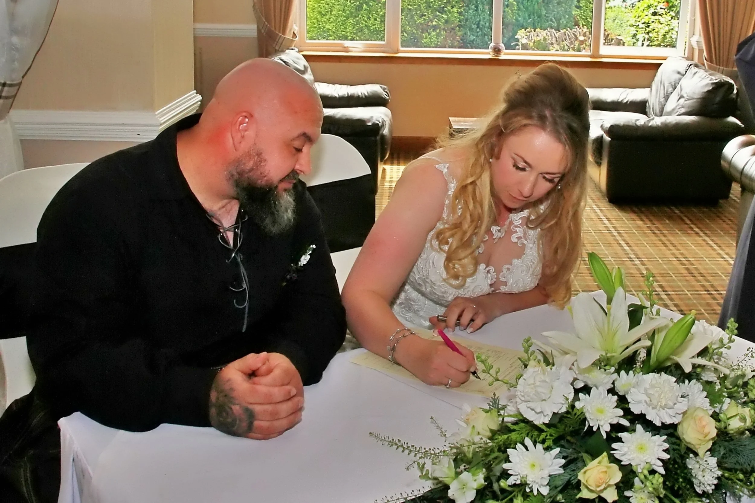 A bride and groom signing a marriage register at a wedding reception, with a floral arrangement in front of them, in a room with large windows and sofas in the background.