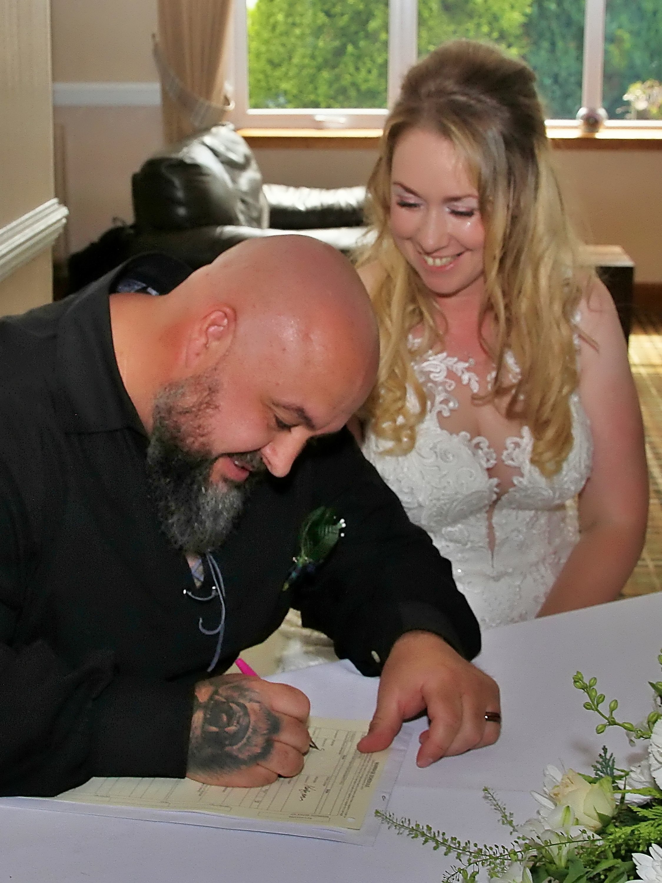 A man with a bald head and tattoos on his hand signs a document on a table, while a woman in a white lace dress looks on and smiles.