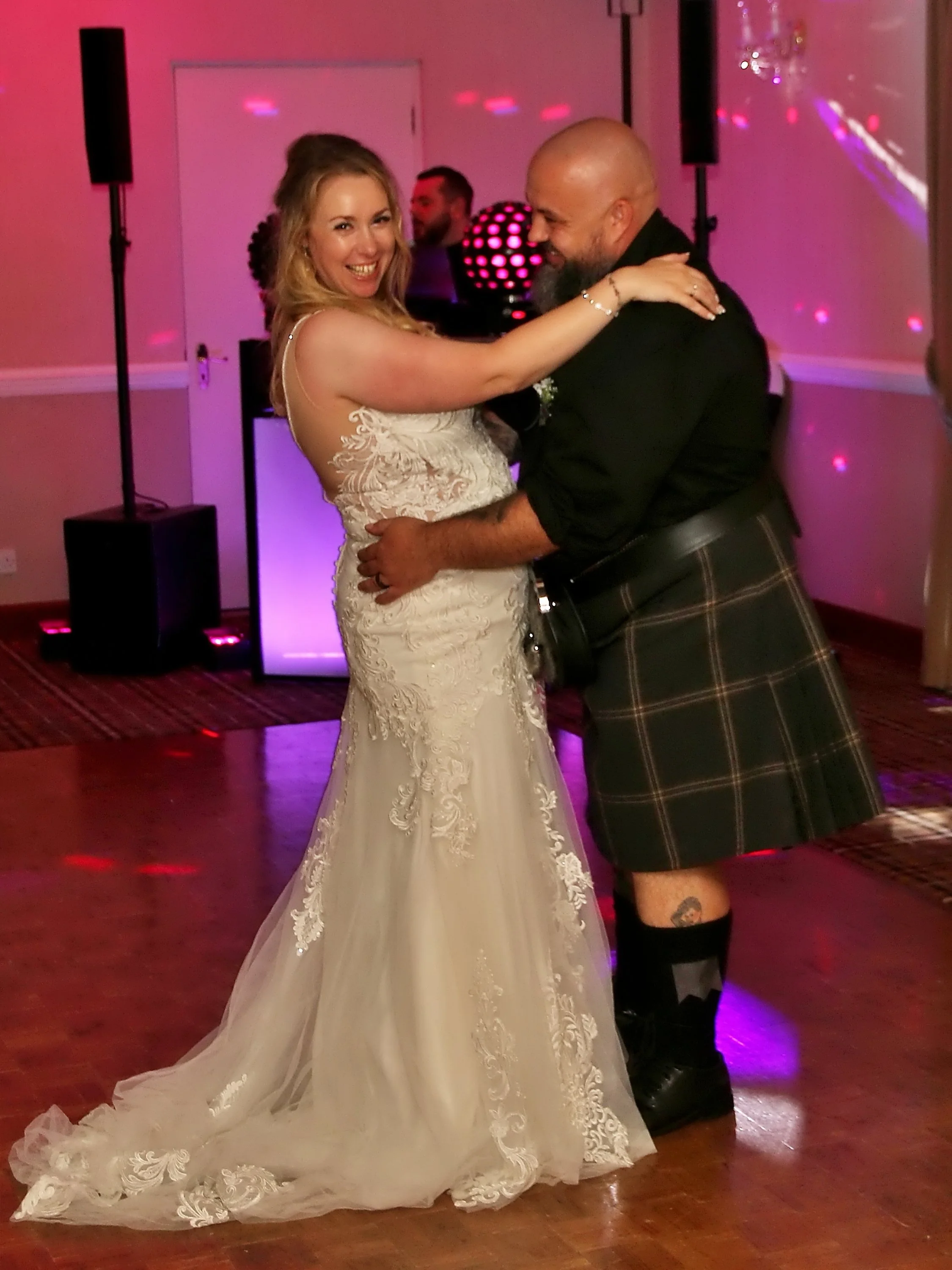 A bride in a white lace wedding dress and a groom in a kilt dancing together at a wedding reception, smiling and looking happy.