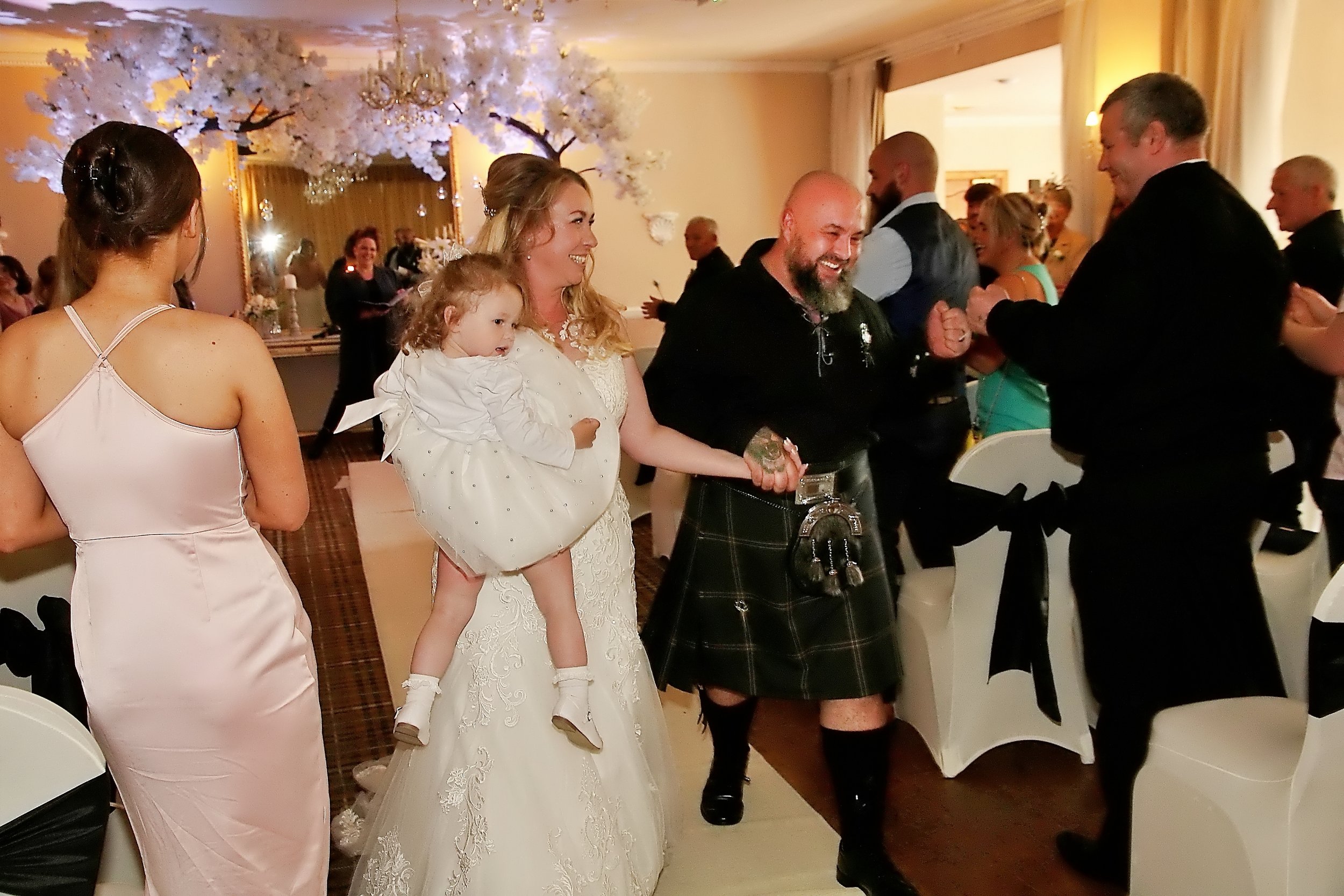 People celebrating at a wedding reception, including a woman in a wedding dress holding a young girl, a man in a kilt, and others dancing and socializing indoors decorated with floral arrangements.