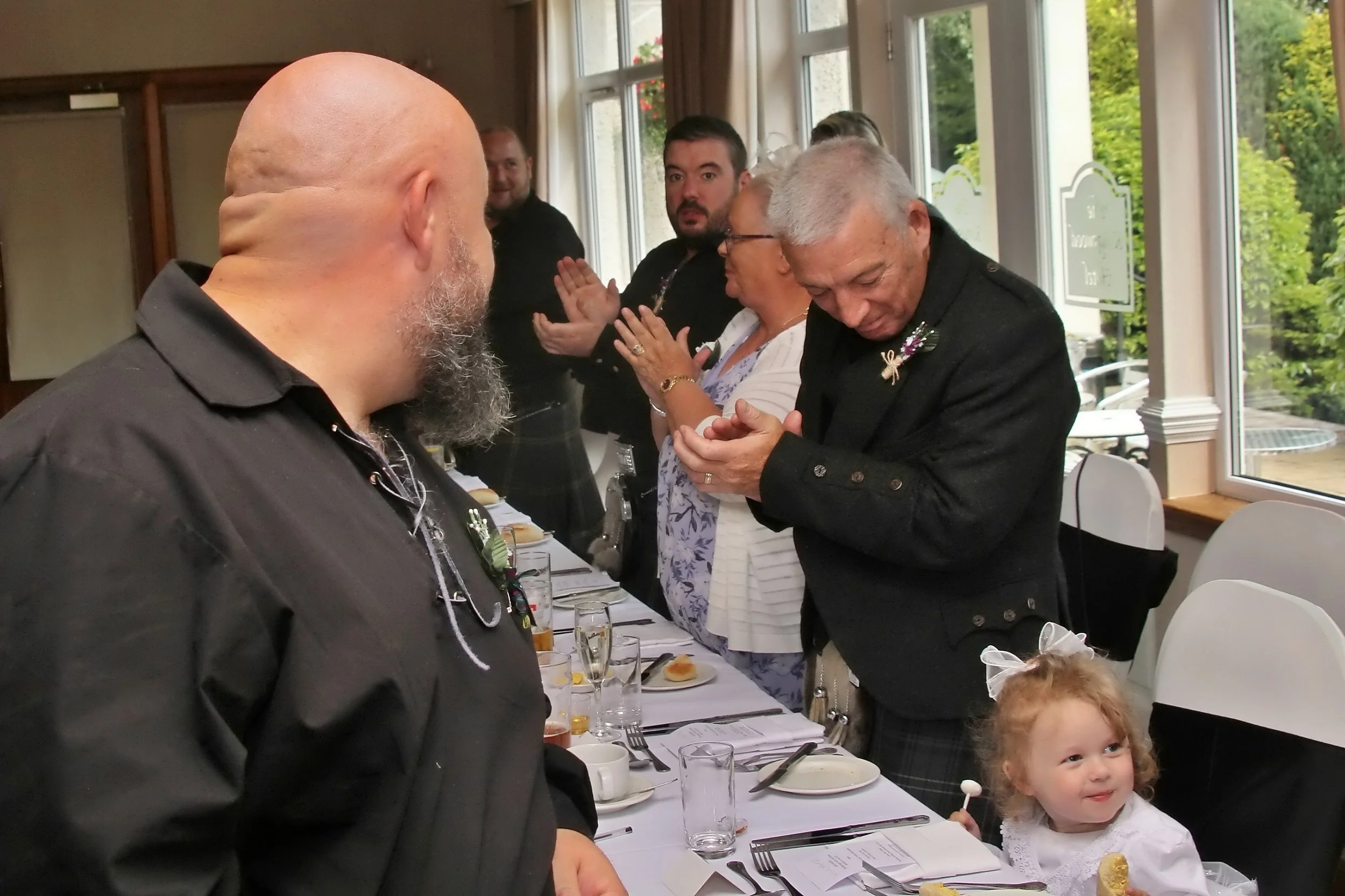 People standing at a dining table during a celebration, with a man in the foreground seen from behind, an elderly man examining a ring, and a young girl with red hair smiling.