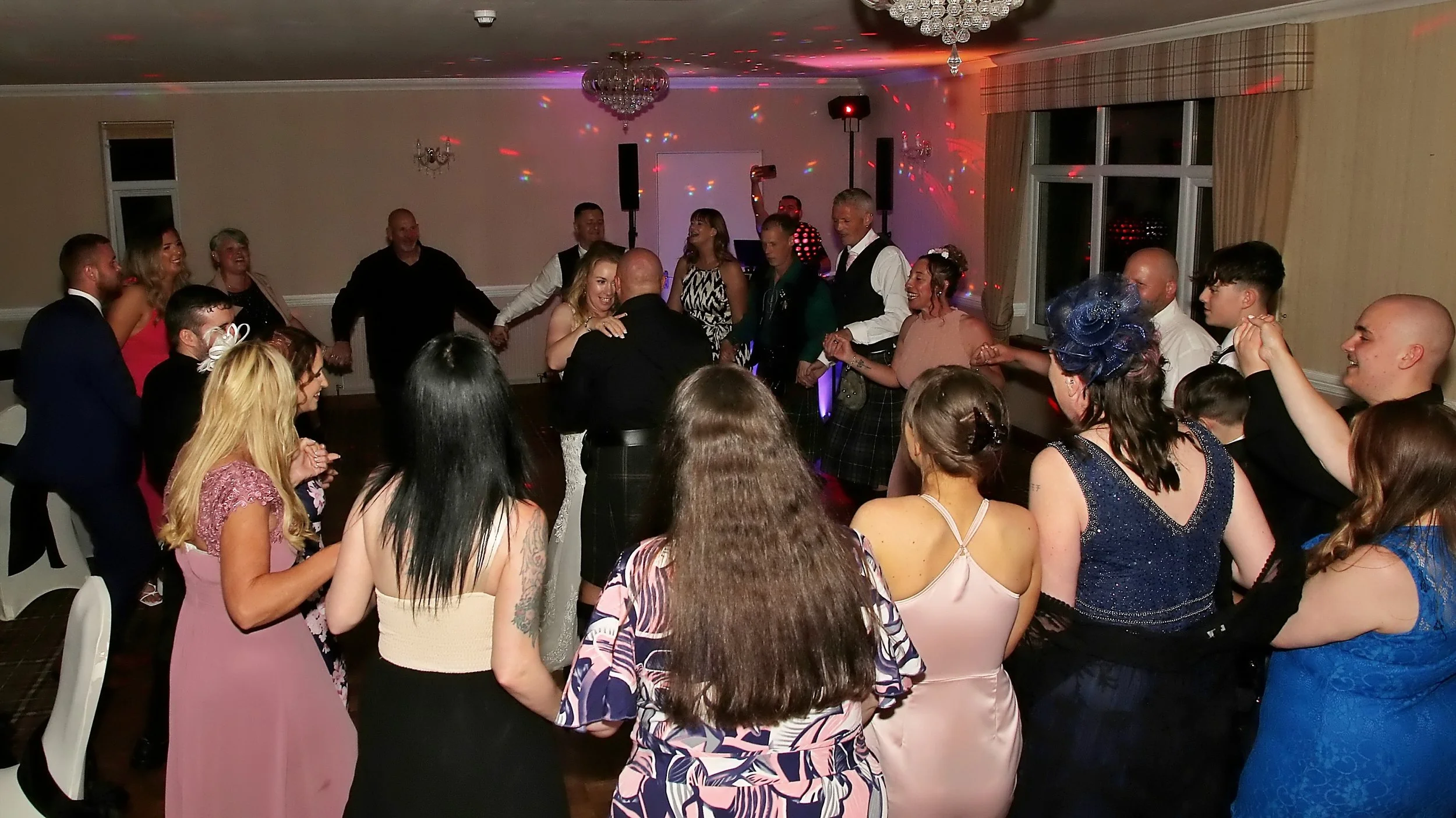 People dancing and celebrating in a decorated reception hall with colorful lights, chandeliers, and windows at night.