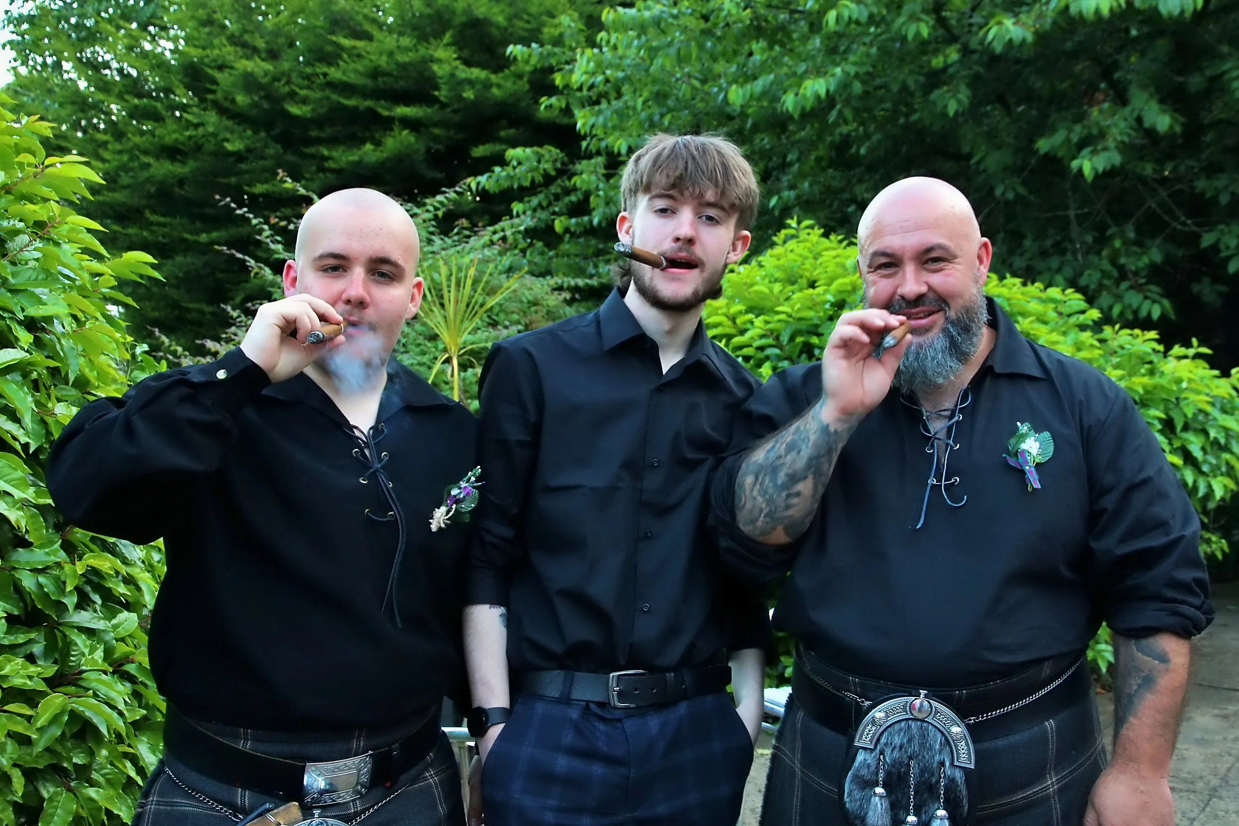 Three men outdoors smoking cigars, dressed in black shirts, with greenery in the background.