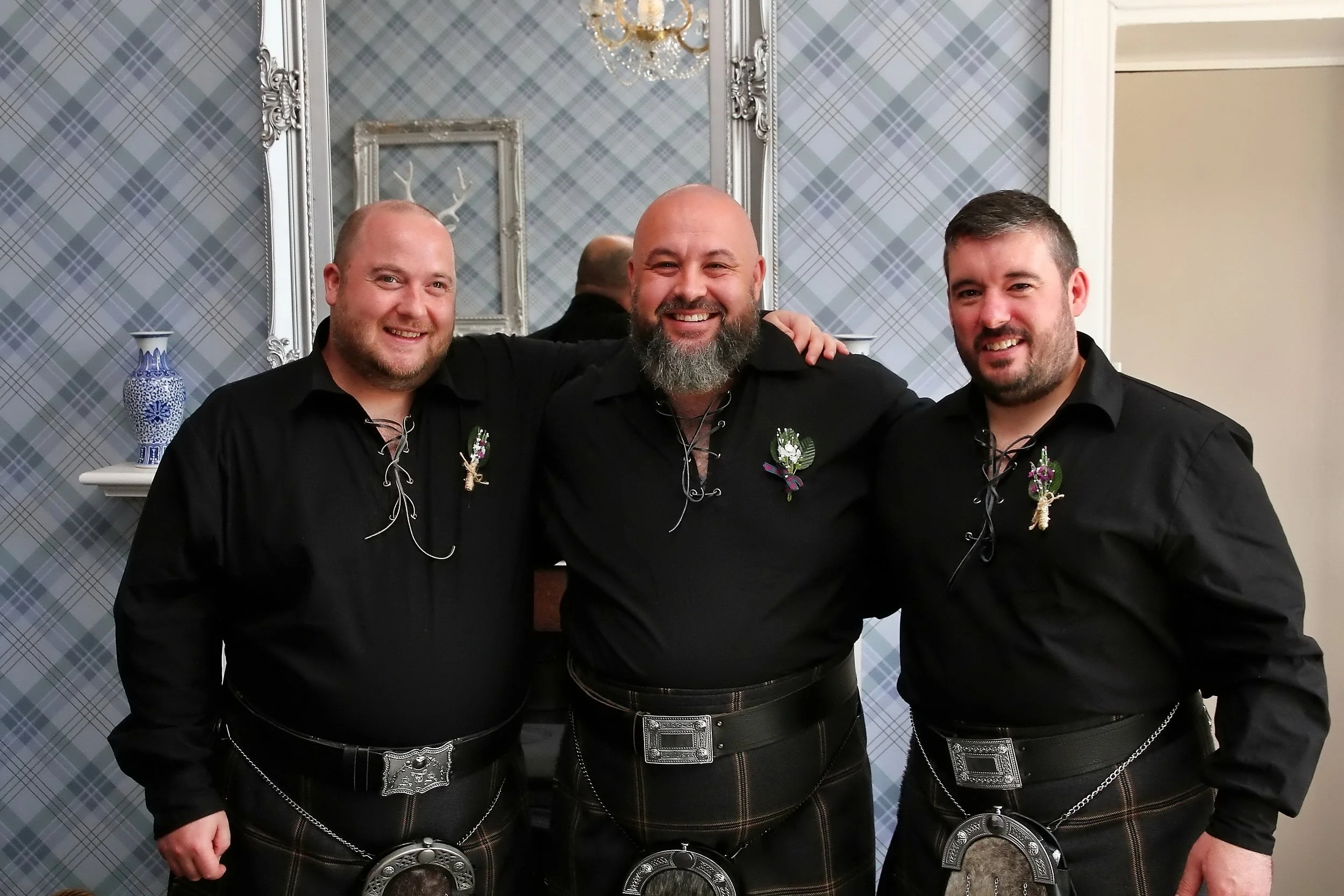 Three men in black shirts and traditional Scottish kilts standing side by side indoors, smiling and posing for the photo.