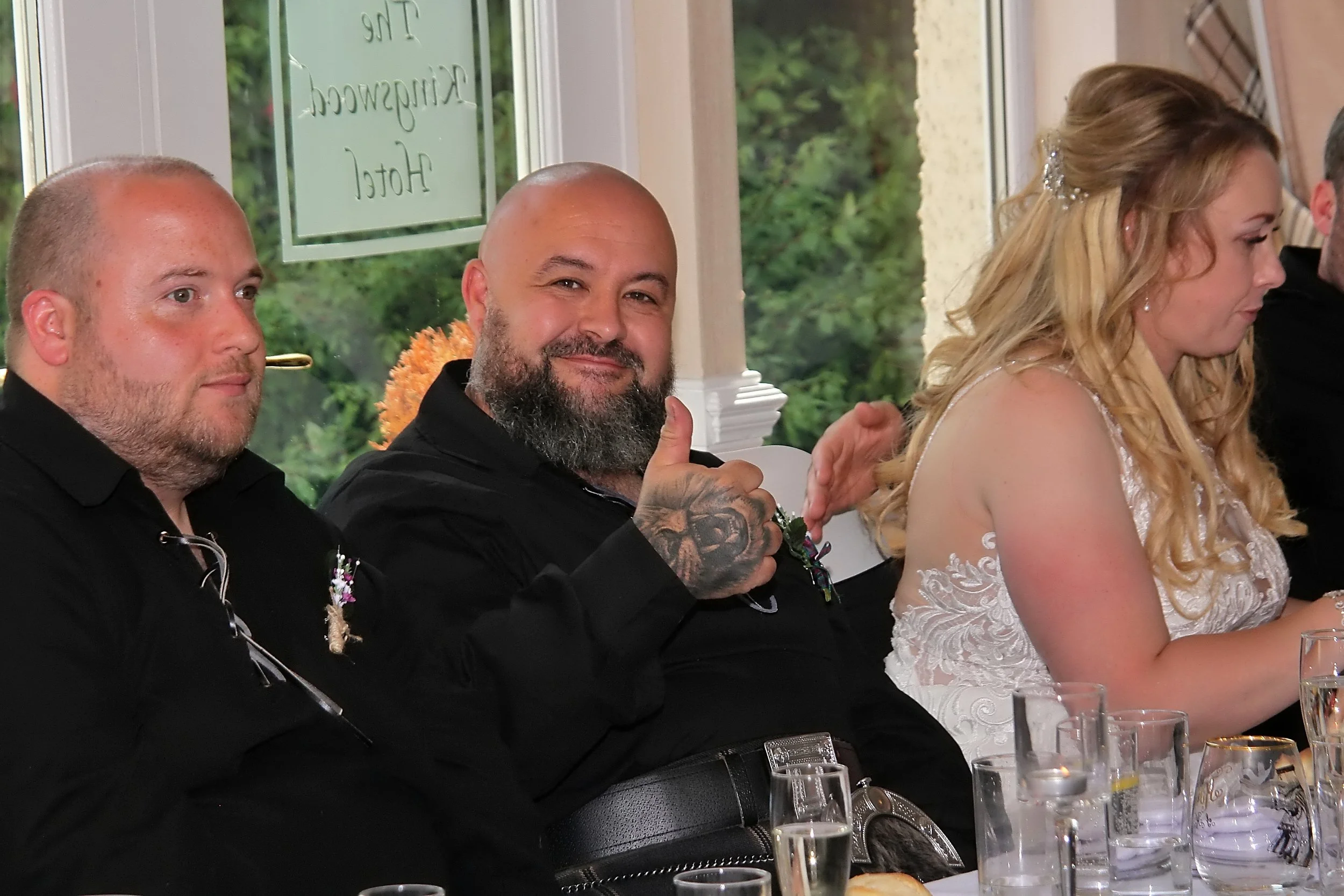 Group of people sitting at a table during a celebration or gathering, with a window in the background showing greenery. One man with a beard is giving a thumbs-up and smiling, while others are looking away or engaged.