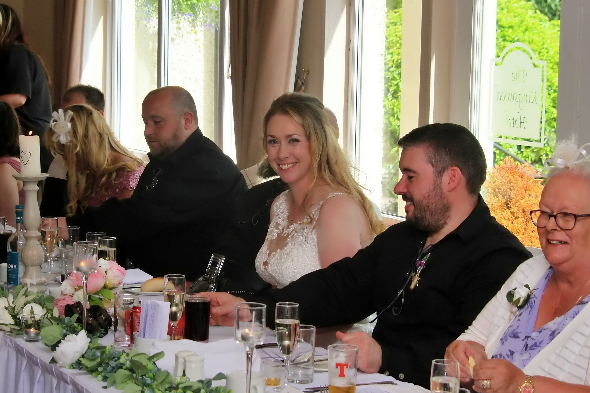 A group of people sitting at a wedding reception table, including a woman in a white lace dress smiling, and others engaged in conversation, with floral centerpieces and drinks on the table.