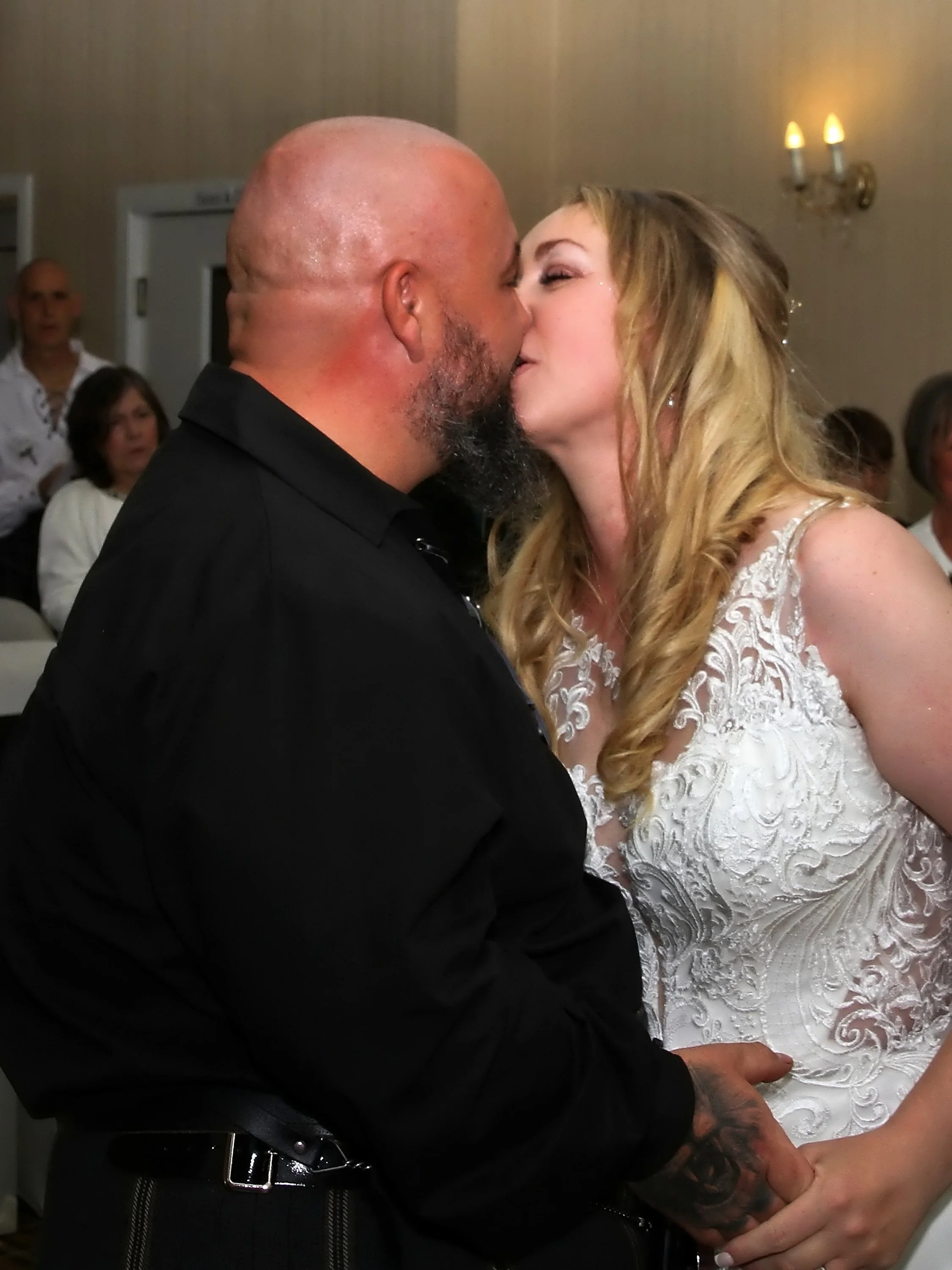 A man and woman kissing at a wedding reception, with guests in the background.