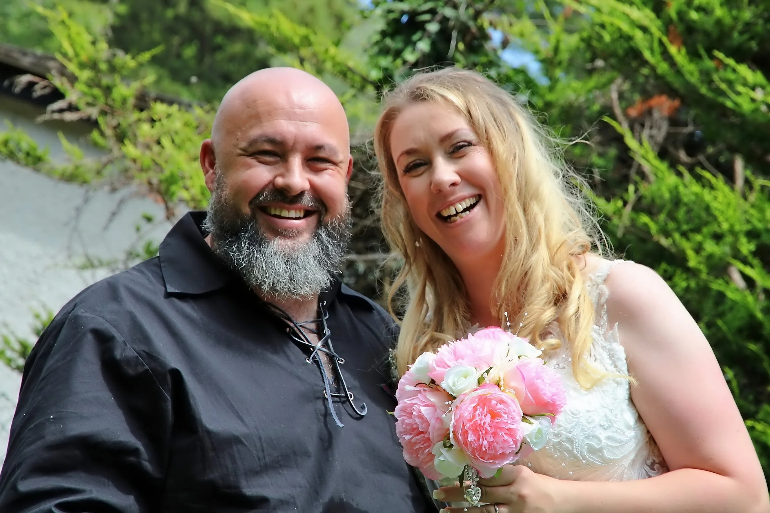 A smiling man with a beard wearing a black shirt, and a smiling woman with blonde hair wearing a white dress, holding a bouquet of pink and white flowers outdoors surrounded by green trees.