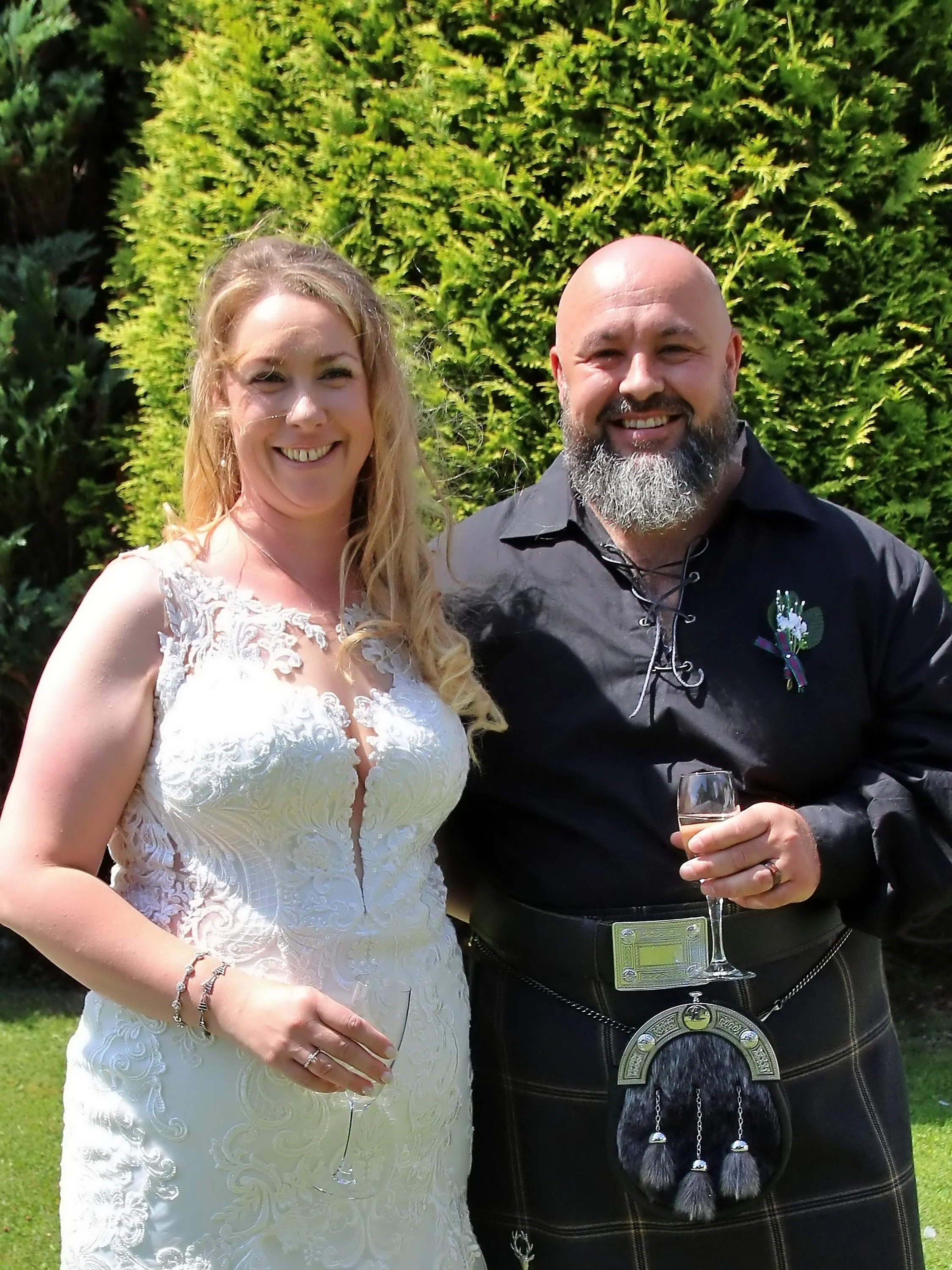 A woman in a white lace wedding dress stands next to a man in traditional Scottish attire, both smiling and holding glasses of champagne, outdoors in front of green shrubbery.
