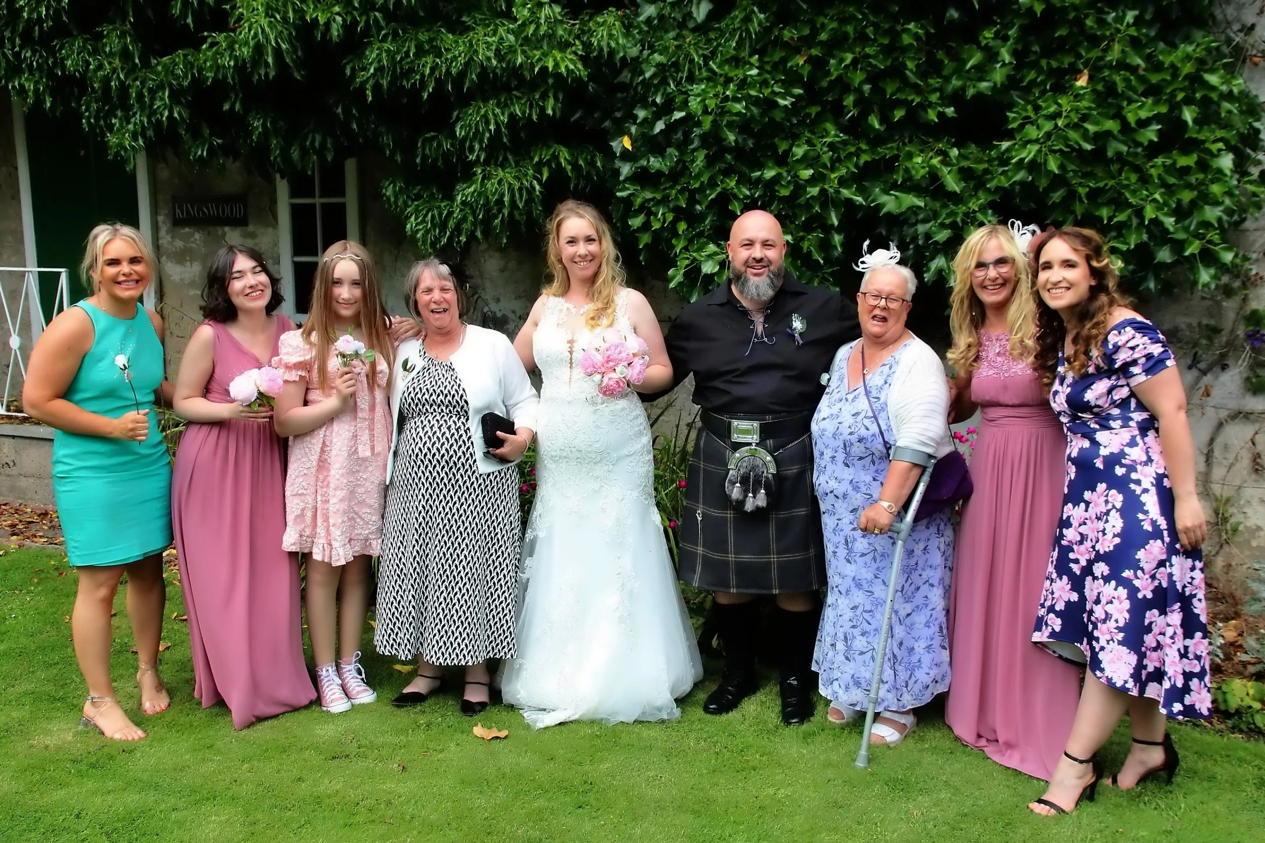 A group of eleven people, including a bride in a white wedding dress holding a pink bouquet, and a groom in a black shirt and kilt, standing on a lawn with a green hedge background, celebrating at a wedding.