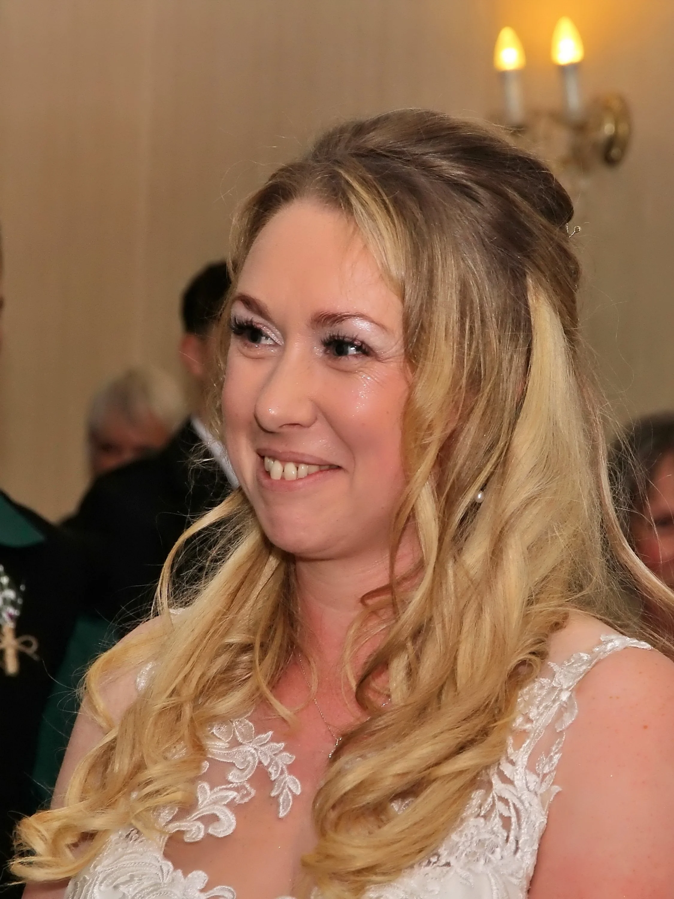 A woman with long, wavy blonde hair smiling, wearing a white lace dress at a formal event.