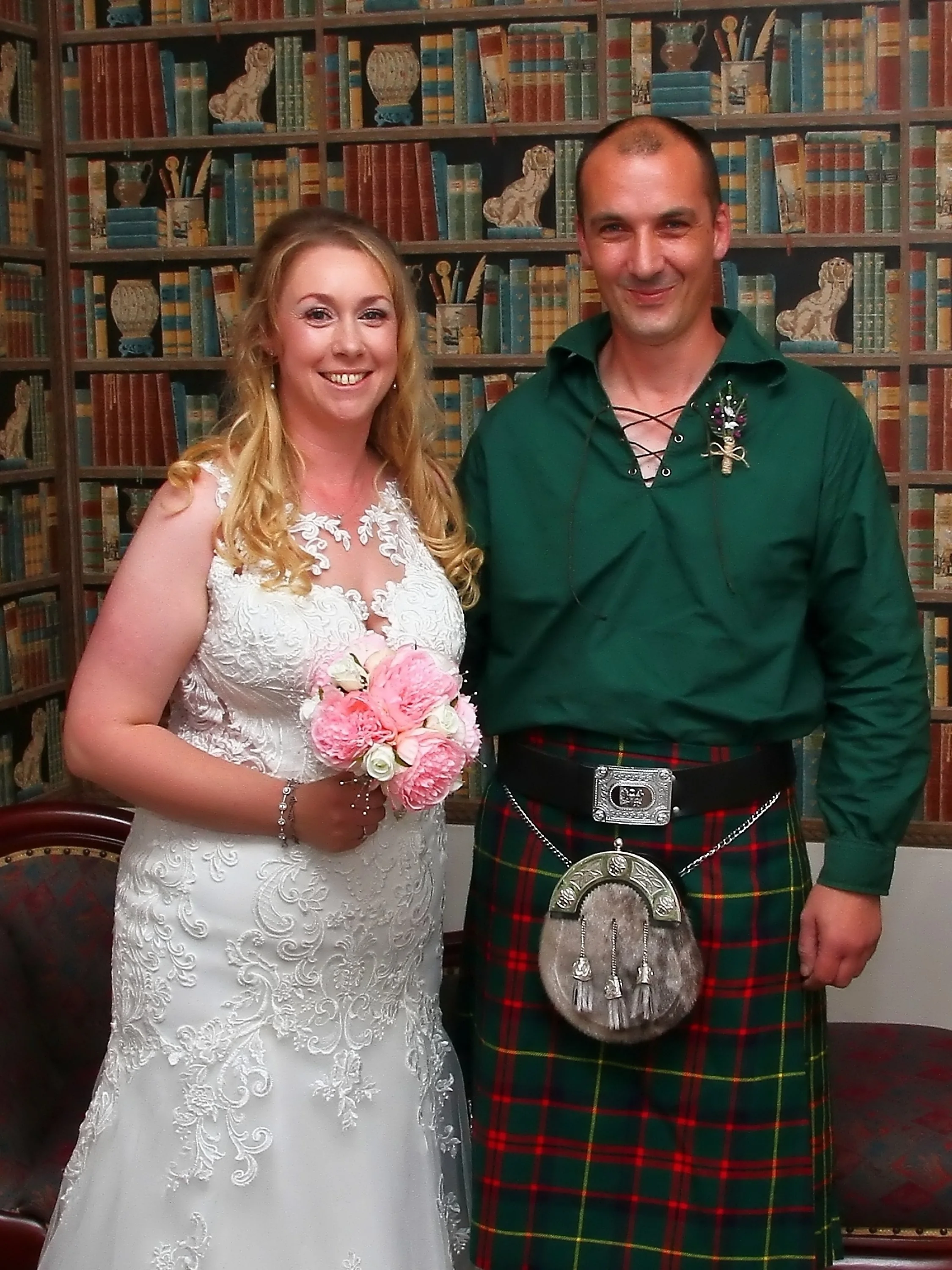 A bride holding a bouquet of pink and white flowers standing next to a man dressed in traditional Scottish attire, including a green kilt and a sporran, in front of a bookshelf background.
