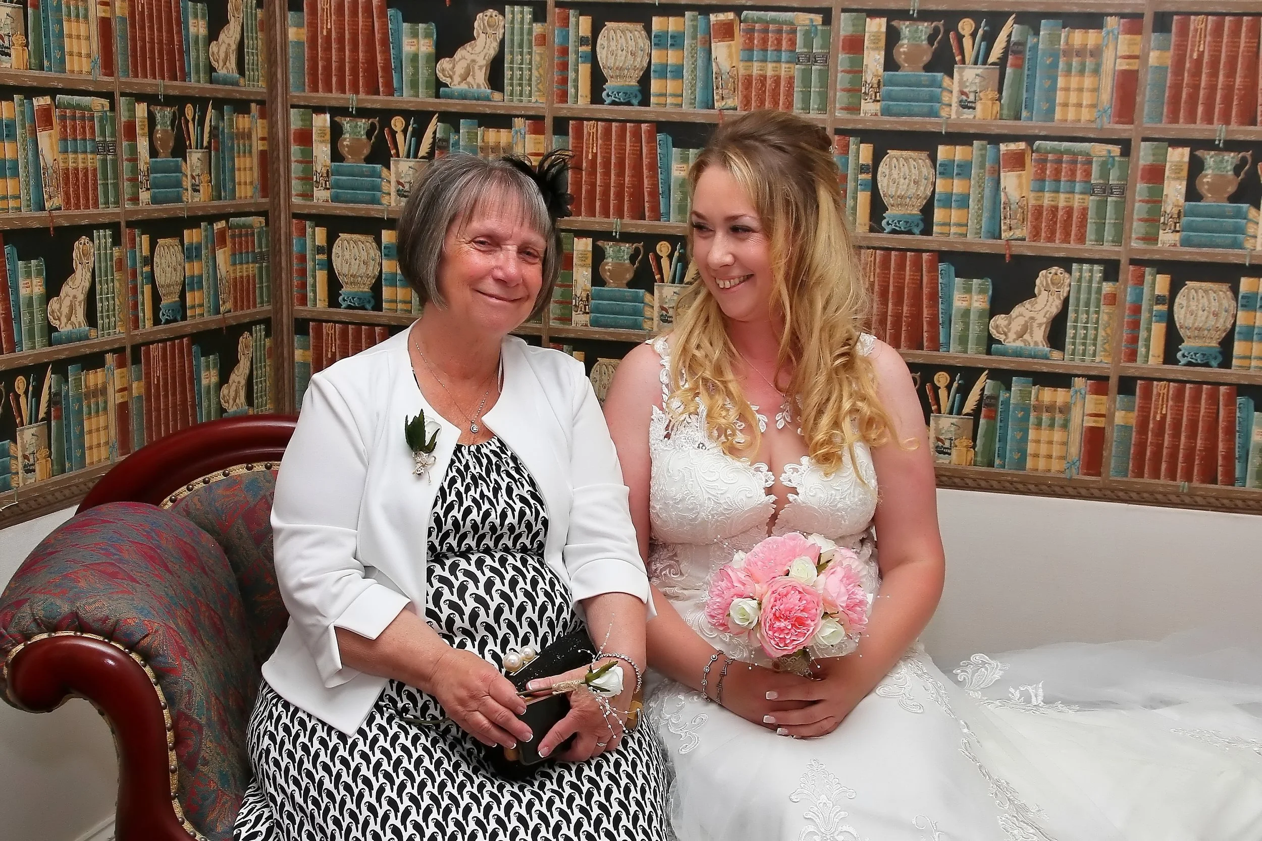 A woman in a white blazer and black-and-white dress sitting next to a young woman in a white wedding dress holding a pink and white bouquet inside a room with a bookcase background.